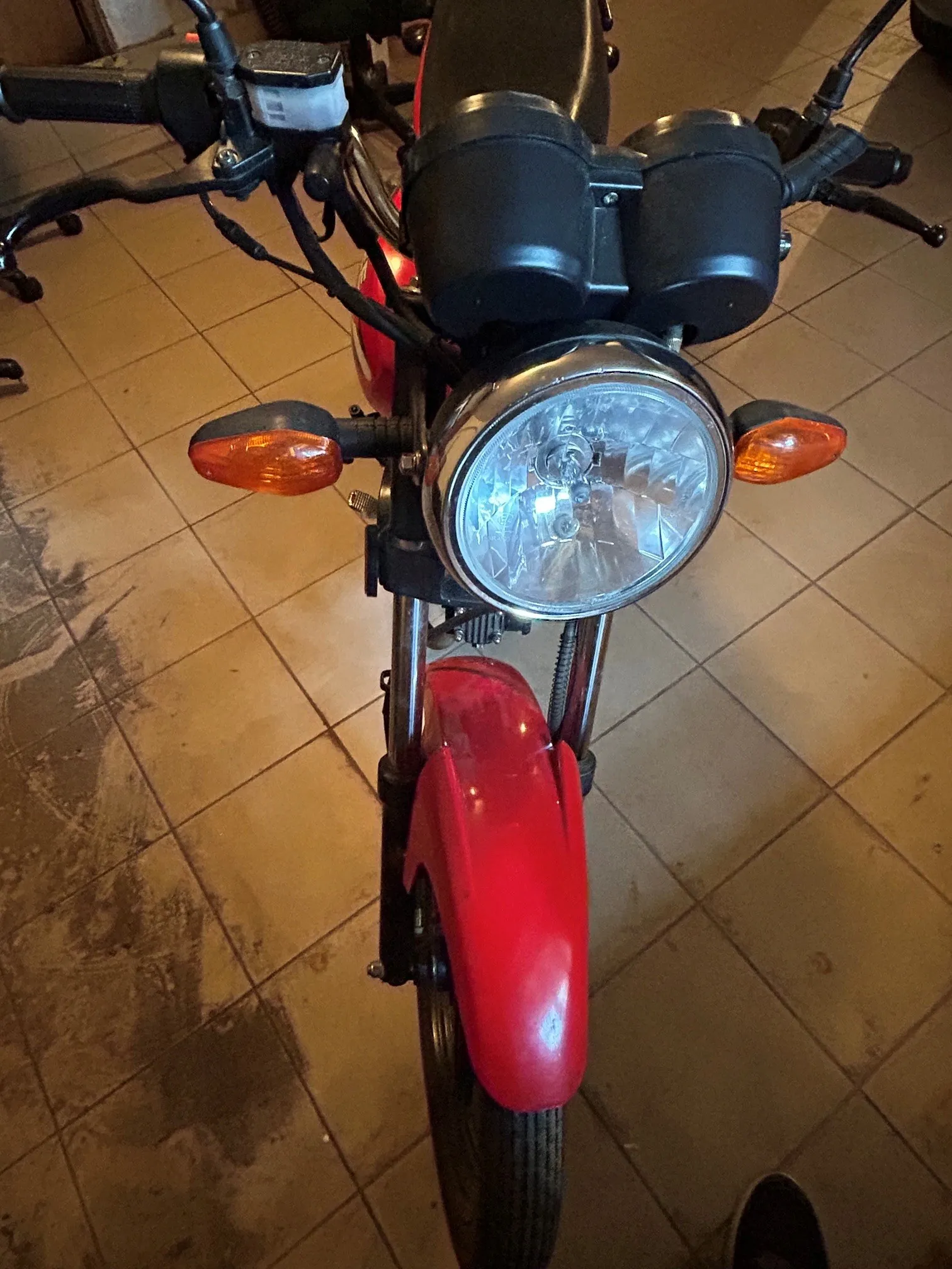 Widok z przodu czerwonego motocykla zaparkowanego na kafelkowej podłodze. Widoczny jest okrągły reflektor główny, dwa pomarańczowe kierunkowskazy po bokach, czarne liczniki nad lampą oraz czerwony błotnik przedni.