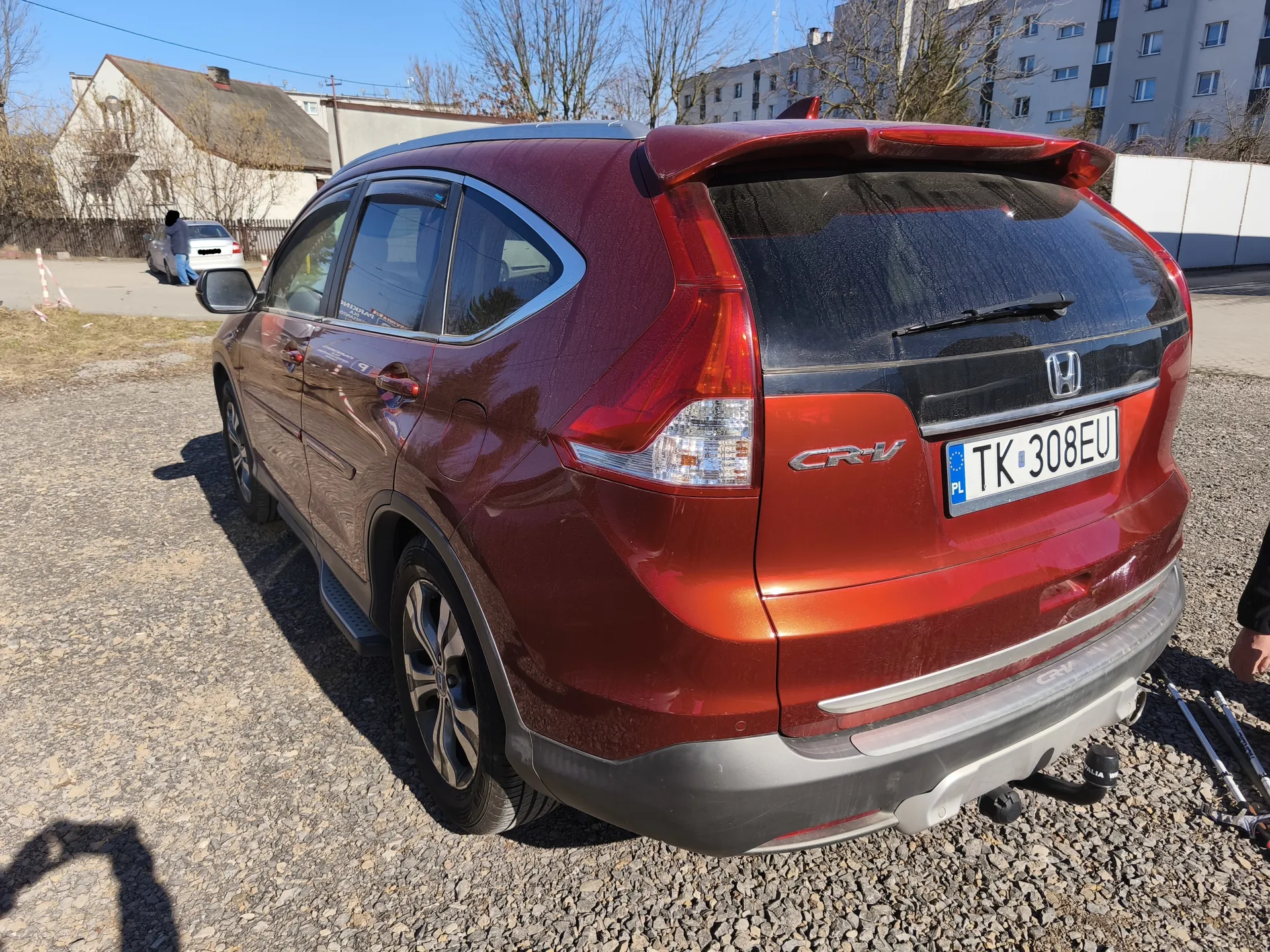 Zdjęcie przedstawia czerwony samochód osobowy marki Honda CR-V zaparkowany na żwirowym podłożu. Widok z tyłu, z widoczną tablicą rejestracyjną PL TK 308EU. W tle budynki mieszkalne, drzewa bez liści, biały samochód oraz osoba stojąca obok niego. Niebo jest bezchmurne. Na tylnym zderzaku widoczny napis 'CR-V' oraz logo Hondy.
