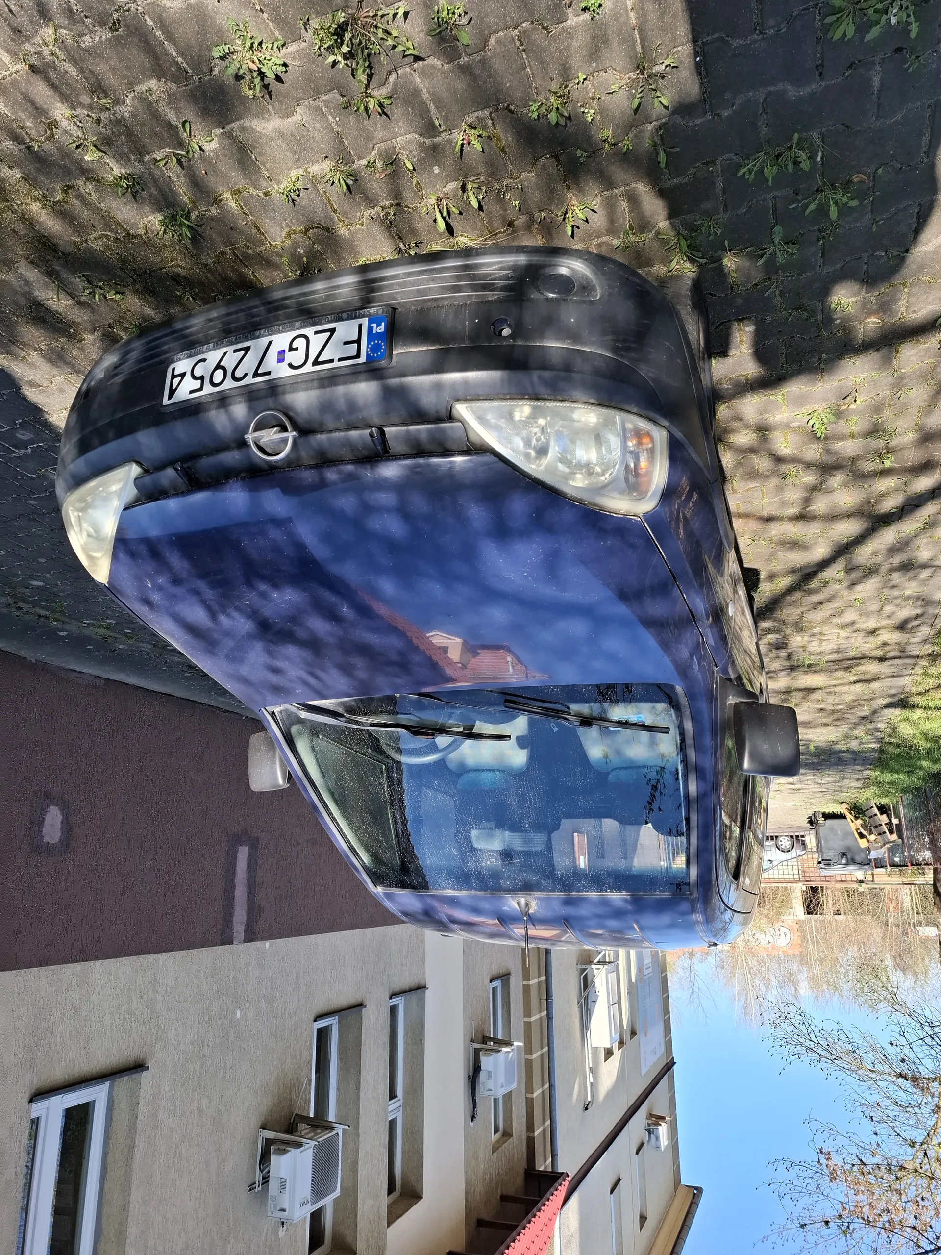 Ciemnoniebieski samochód marki Opel zaparkowany na brukowanej powierzchni. Widoczna tablica rejestracyjna FZG 72954 z kodem kraju PL. Po lewej stronie budynki z oknami i klimatyzatorami. Jasne niebo, drzewa oraz inne budynki w tle. Na przedniej szybie refleksy, a na bruku rosną pojedyncze rośliny.