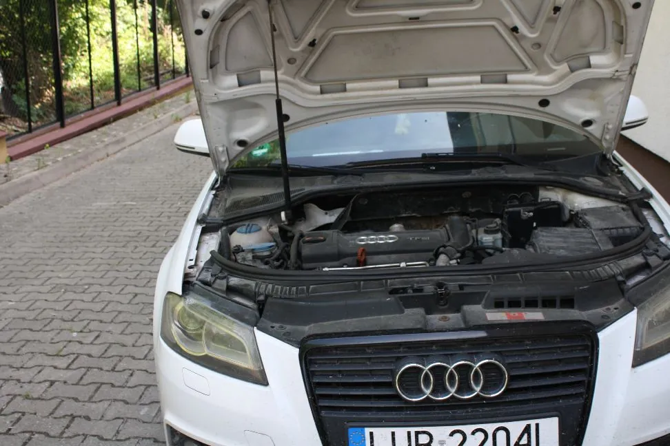 Widok z przodu białego samochodu marki Audi z otwartą maską, odsłaniającą silnik. Pojazd stoi na kostce brukowej obok czarnego ogrodzenia. Na grillu widoczne jest logo Audi, a poniżej fragment tablicy rejestracyjnej z tekstem 'LUB 2204L'.