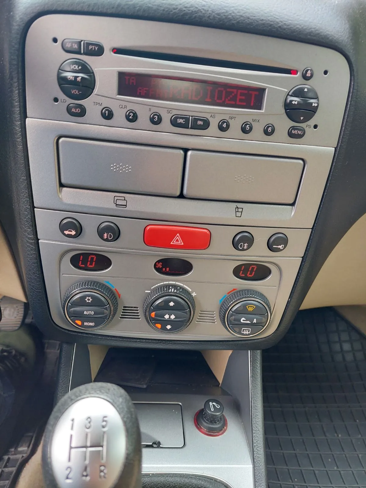 Zdjęcie wnętrza samochodu przedstawiające konsolę środkową. Na górze znajduje się radio z wyświetlaczem LCD, na którym widnieje napis 'RADIOZET'. Poniżej znajdują się dwa schowki, czerwony przycisk alarmowy oraz panel sterowania klimatyzacją z dwoma cyfrowymi wyświetlaczami pokazującymi wartość 'LO' i trzema pokrętłami. Na dole widoczna jest lewarek zmiany biegów z oznaczeniami biegów 1-6 oraz wstecznego (R).