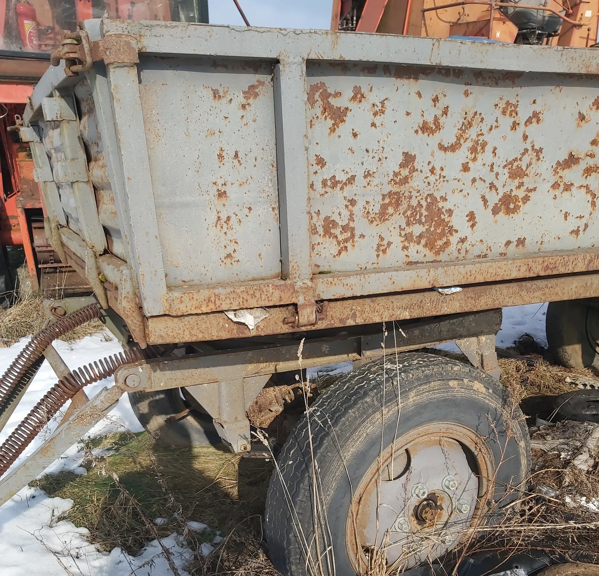 Zardzewiały metalowy przyczepa lub zbiornik o szarym kolorze z brązowymi plamami rdzy. Widoczne są metalowe panele, zardzewiałe zawiasy i zatrzaski po lewej stronie. Przyczepa jest zamocowana do pomarańczowego pojazdu (prawdopodobnie ciągnika) w tle. Na dole obrazu znajdują się duże czarne opony z metalowymi felgami, częściowo zakryte suchą trawą i śniegiem. W tle widoczne są również pomarańczowe elementy maszyn oraz fragment nieba. Na ziemi śnieg i sucha trawa.