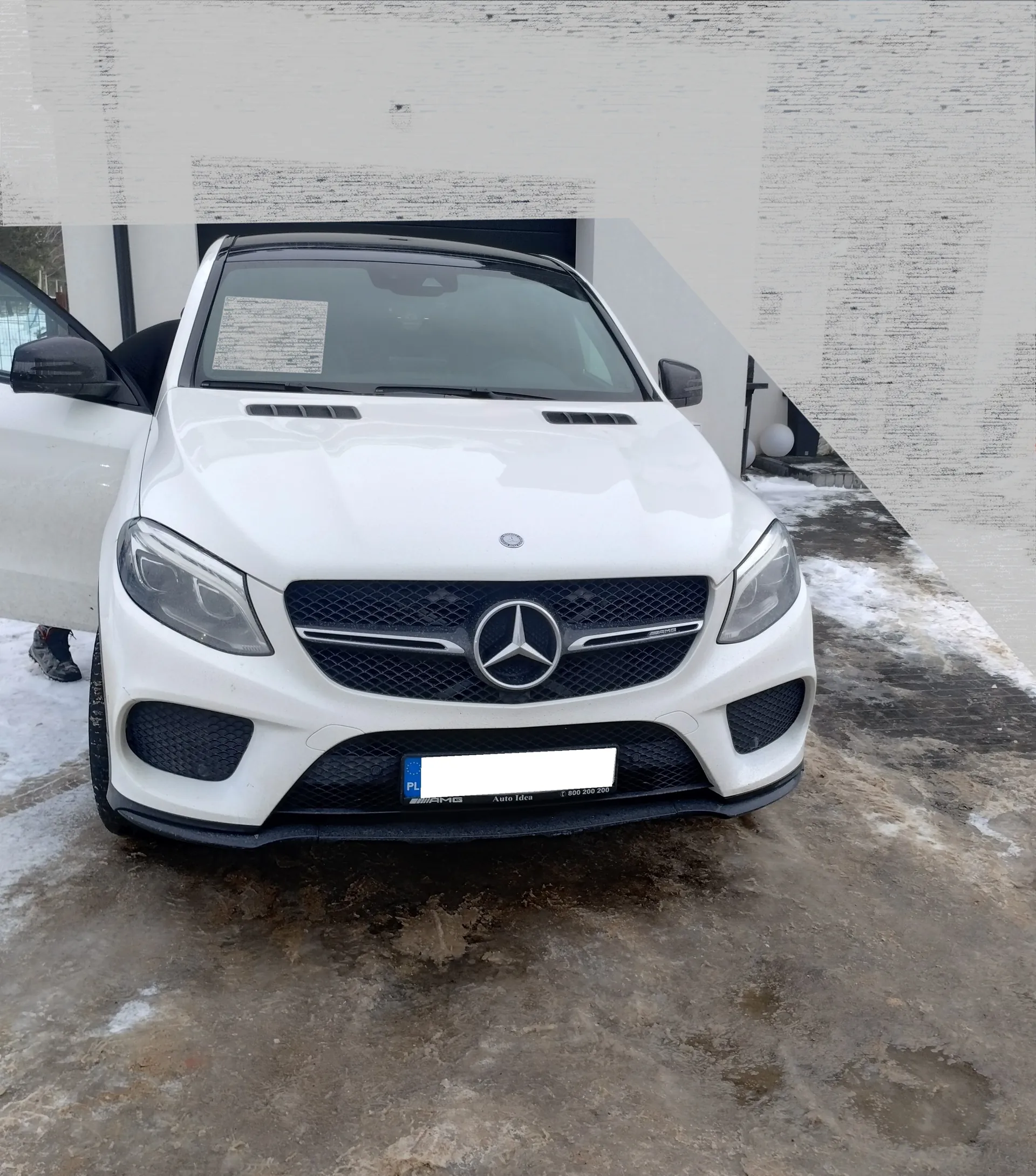 Biały samochód osobowy marki Mercedes-Benz z widocznym logo trójłabędzia na atrapie chłodnicy. Przedni widok, otwarte drzwi kierowcy. Tablica rejestracyjna z oznaczeniem 'PL', napisem 'Auto Idea' oraz numerem telefonu '800 200 200'. Na tle budynek z białą ścianą i śniegiem na ziemi. Obok otwartych drzwi widoczny but. Na przedniej szybie dokument z tekstem.