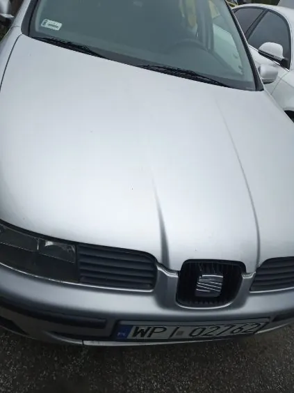Przód srebrnego samochodu marki SEAT z widocznym logo na masce. Tablica rejestracyjna z napisem WPJ 02762 umieszczona na przednim zderzaku. W tle częściowo widoczny biały samochód. Samochód zaparkowany na asfaltowej powierzchni.