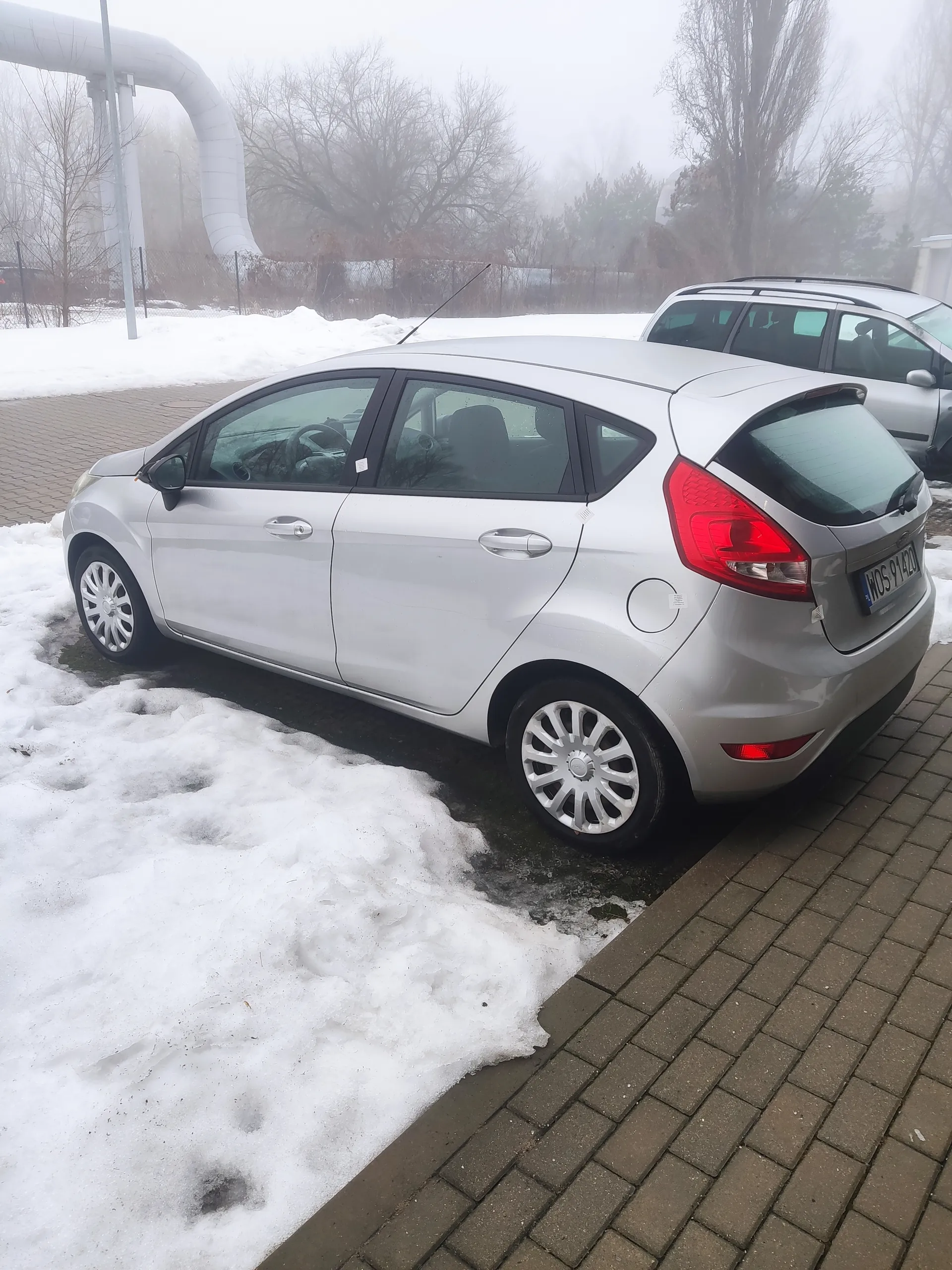 Srebrny samochód osobowy typu hatchback zaparkowany obok ceglanej ściany. Na ziemi znajduje się śnieg, w tle mgła, drzewa bez liści oraz biały rurociąg. Widoczne są koła z aluminiowymi felgami i czerwone światła tylne.