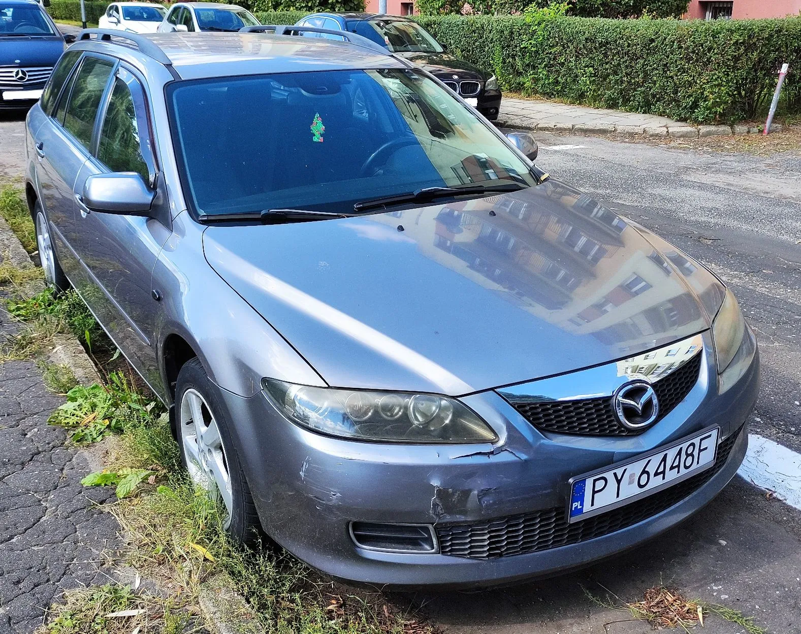 Srebrnoszary samochód marki Mazda w nadwoziu kombi zaparkowany przy krawężniku na ulicy. Pojazd ma widoczne uszkodzenia w postaci wgnieceń i zarysowań na przednim zderzaku po prawej stronie. Tablica rejestracyjna ma numer PY 6448F. W tle widoczne są inne zaparkowane samochody oraz zielony żywopłot.