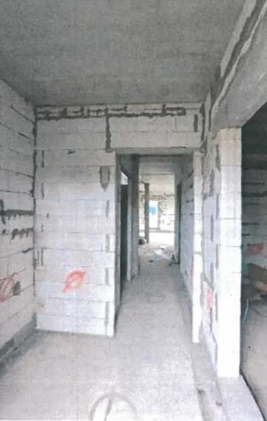 Wnętrze budynku w trakcie budowy z betonowymi blokami tworzącymi ściany korytarza. Widoczne są otwarte przejścia prowadzące do kolejnych pomieszczeń, betonowa podłoga i sufit. Na ścianach znajdują się czerwone znaczniki lub napisy. Oświetlenie jest przygaszone, a przestrzeń wydaje się nieukończona.