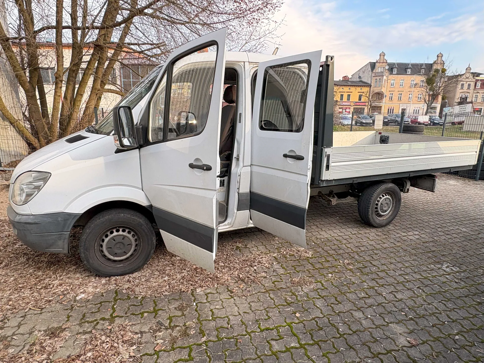 Biały samochód dostawczy z otwartymi drzwiami przodem i bokiem, z platformą załadowczą. Pojazd stoi na brukowanej powierzchni z suchymi liśćmi. W tle drzewa bez liści, budynki mieszkalne i handlowe oraz niebo z chmurami.