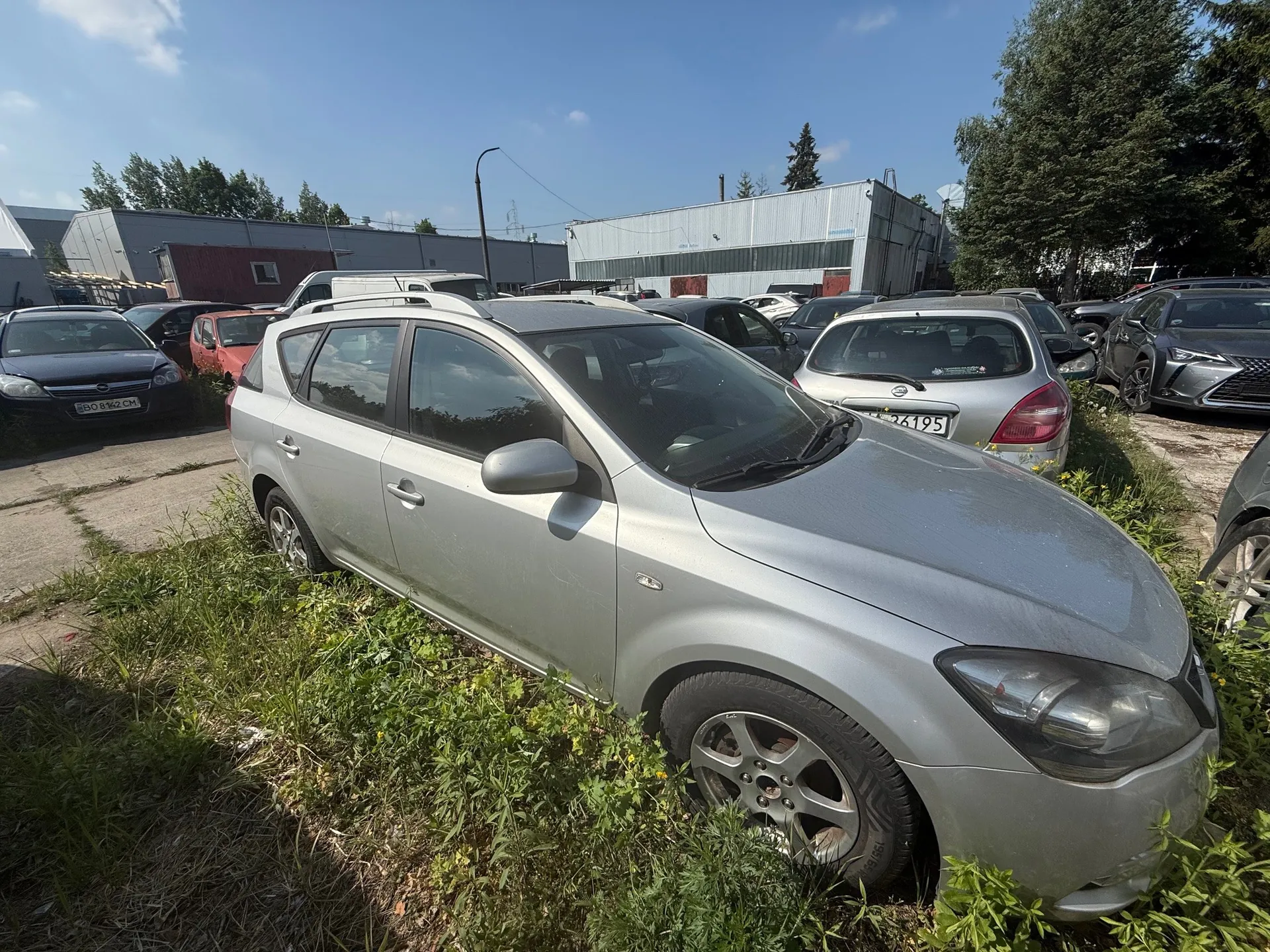 Srebrny samochód typu kombi zaparkowany na porośniętym trawą obszarze. W tle inne pojazdy, w tym czarny Opel z tablicą rejestracyjną BO 8142 CM oraz srebrny Nissan z tablicą 26195. Widoczne są przemysłowe budynki, drzewa i błękitne niebo z kilkoma chmurami.