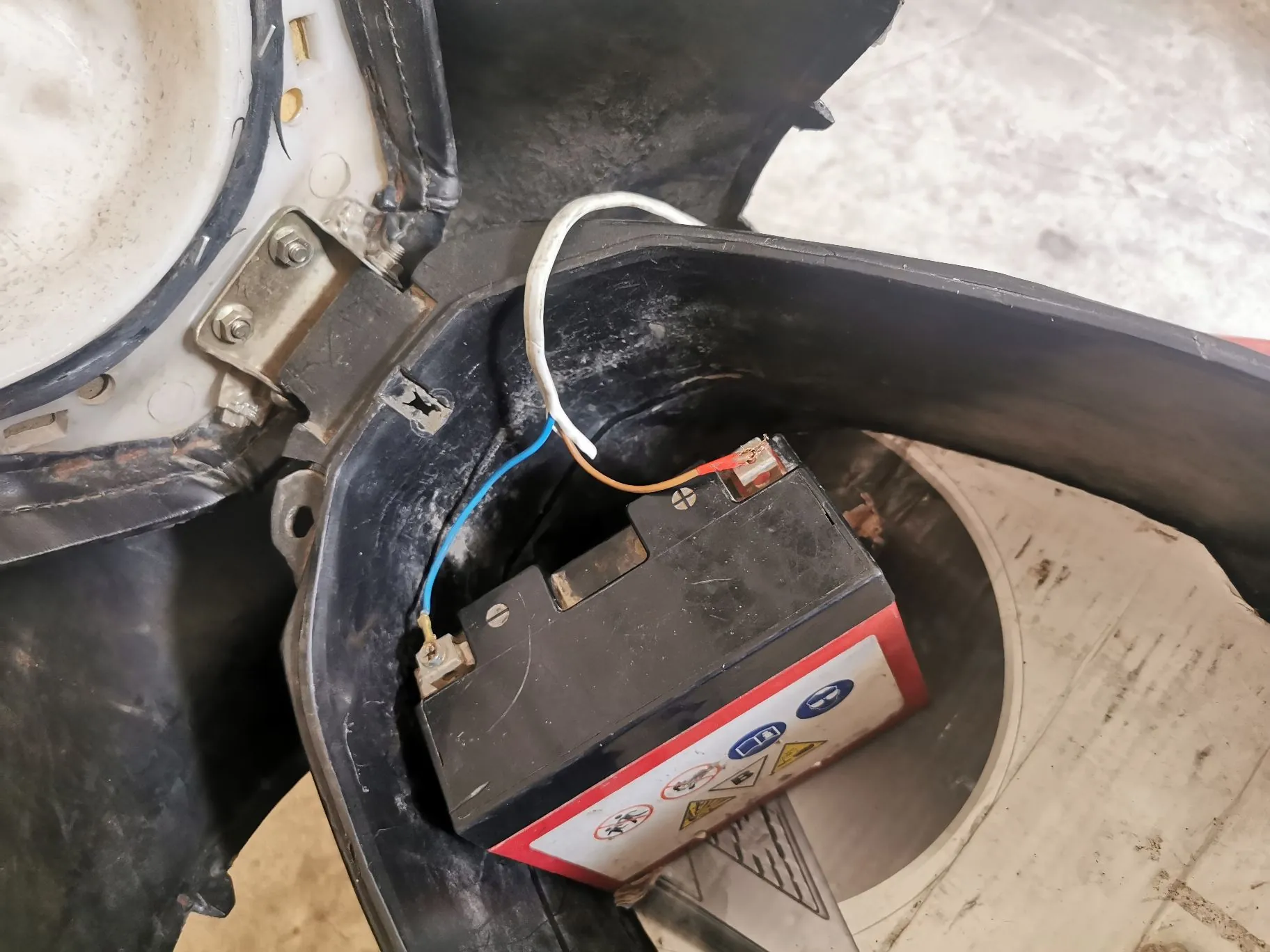 Zbliżenie na czarną baterię akumulatorową umieszczoną w plastikowym schowku pojazdu. Do akumulatora podłączone są przewody w kolorach niebieskim, białym i czerwonym. Na boku akumulatora widoczna jest biała etykieta z czerwonymi i niebieskimi symbolami ostrzegawczymi oraz piktogramami.