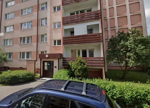 Wielopiętrowy blok mieszkalny w kolorach beżowym i różowym z białymi ramami okien i brązowymi balkonami. Przed budynkiem znajduje się niebieski samochód z bagażnikiem dachowym, zielone krzewy oraz drzewo po prawej stronie.