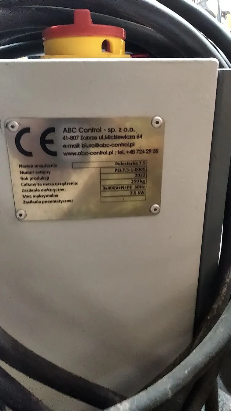 Zbliżenie na białą obudowę urządzenia elektrycznego z przyklejoną tabliczką znamionową. Na tabliczce widnieje logo CE oraz dane firmy: 'ABC Control - sp. z o.o., 41-807 Zabrze ul. Mickiewicza 64, e-mail: biuro@abc-control.pl, www.abc-control.pl; tel. +48 724 29 58'. Tabela na tabliczce zawiera informacje: Nazwa urządzenia: Peleciarka 7.5, Numer seryjny: PEL7,5-1-0005, Rok produkcji: 2022, Całkowita masa urządzenia: 250 kg, Zasilanie elektryczne: 3x400V+N+PE 50Hz, Moc maksymalna: 7,5 kW. Nad obudową widoczny jest żółto-czerwony przycisk bezpieczeństwa oraz czarne kable.