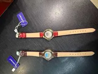 Dwa zegarki na ciemnej powierzchni. Górny zegarek ma czerwone elementy na pasku i niebieską etykietę z białym napisem 'DKNY'. Dolny zegarek ma brązowe elementy na pasku i niebieską etykietę z białym napisem 'DKNY'. Oba zegarki mają jasne paski i okrągłe tarcze.