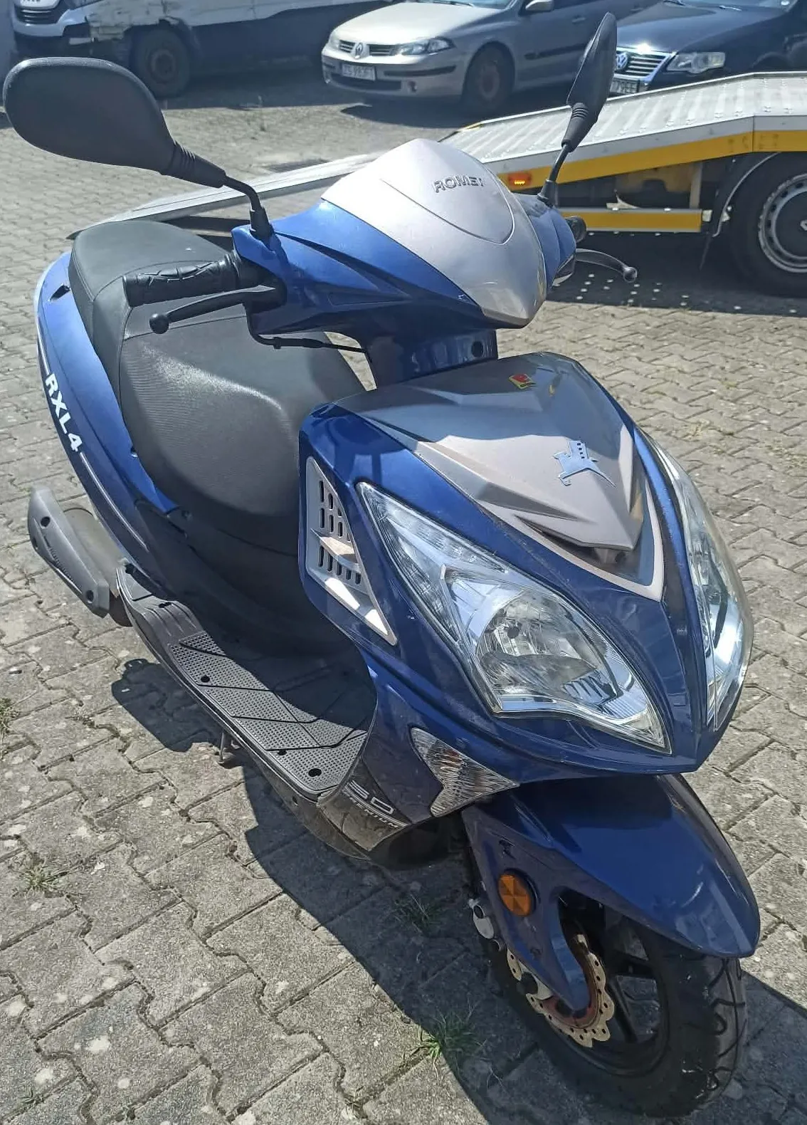 Niebieski skuter marki Romee, model RXL4, zaparkowany na kostce brukowej. Pojazd jest widoczny z przodu pod kątem, z widoczną przednią lampą, lusterkami i siedzeniem. W tle widać fragmenty samochodów oraz żółtą przyczepę.