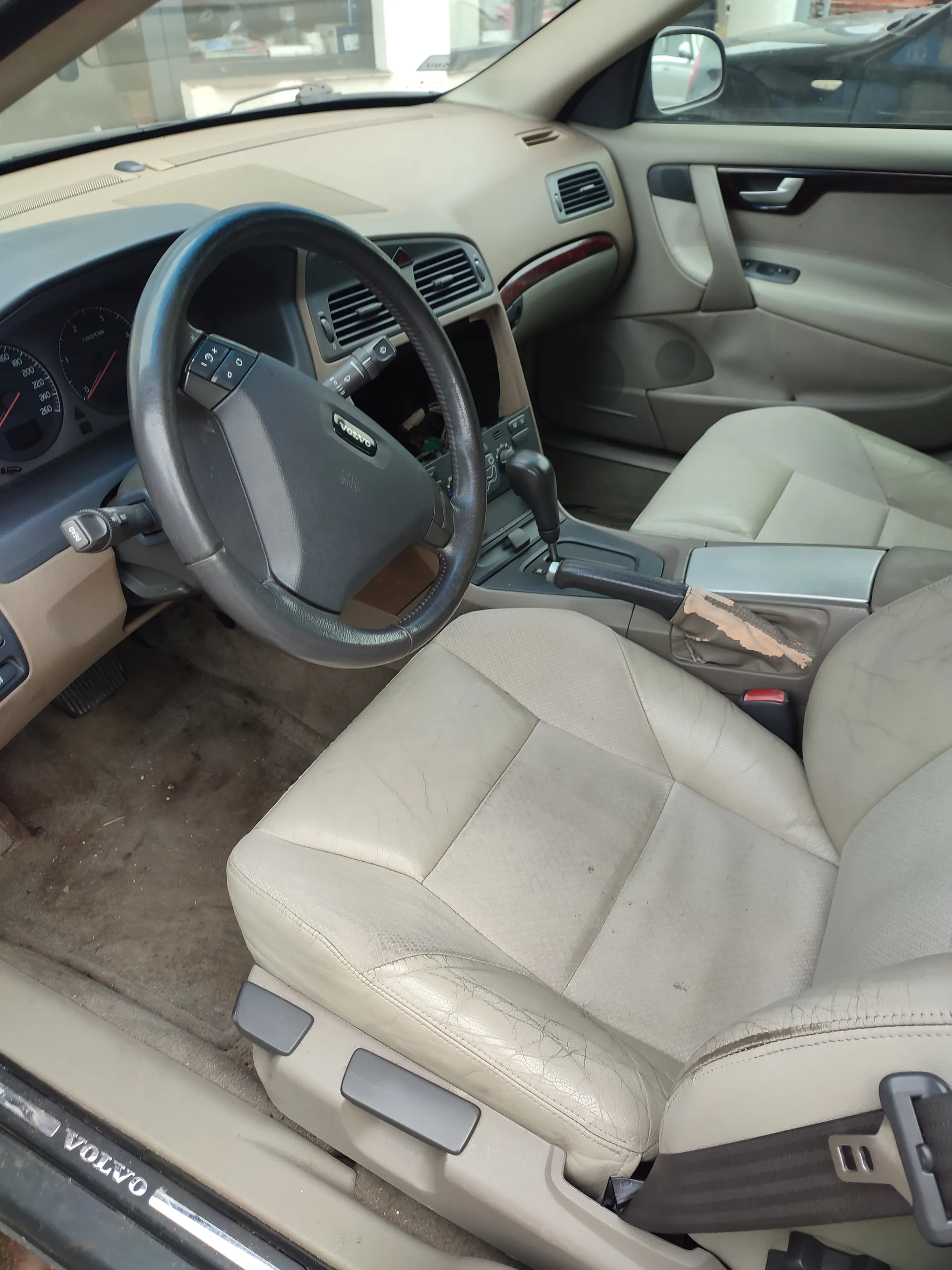 Wnętrze samochodu marki Volvo z beżowymi skórzany siedzeniami, kierownicą z logo Volvo, deską rozdzielczą z licznikami, dźwignią zmiany biegów i otwartą drzwiami. Widoczne są uszkodzenia na siedzeniach oraz na centralnej konsoli. Na progu drzwi widnieje napis VOLVO.