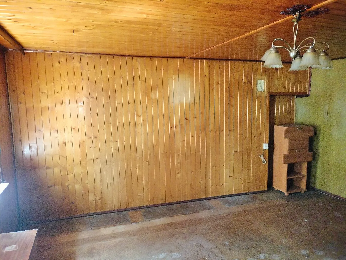 Wnętrze pomieszczenia z drewnianą boazerią na ścianach i suficie. Po prawej stronie znajduje się drewny mebel przypominający organy lub szafkę z półkami. Z sufitu zwisa żyrandol z czterema białymi kloszami. Podłoga jest ciemna i gładka.