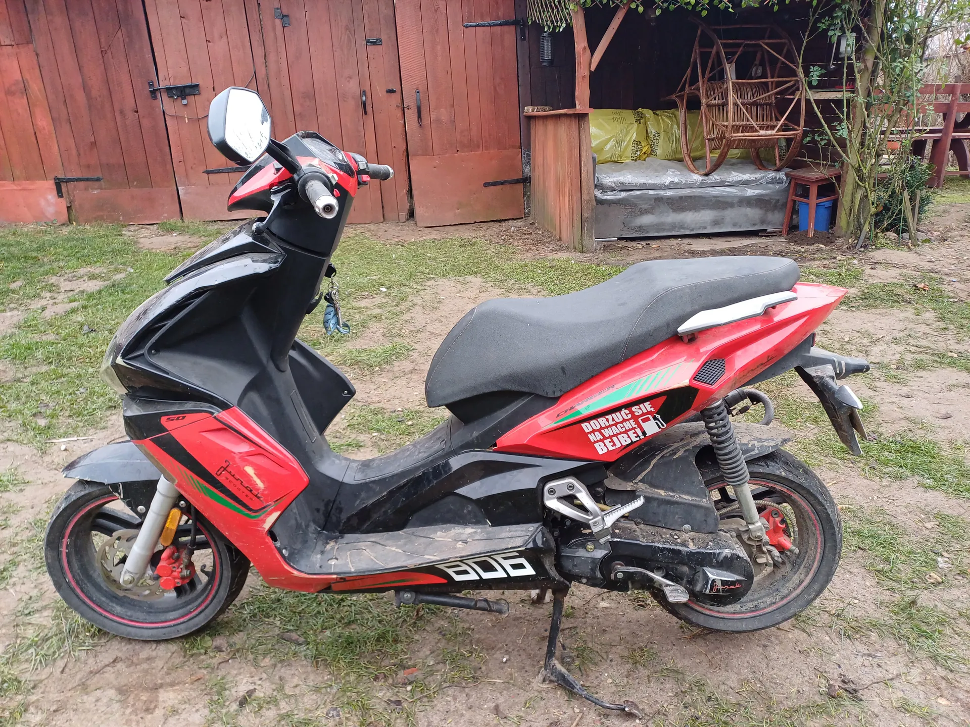 Na zdjęciu czerwono-czarny, zabrudzony skuter z czarnym siedzeniem, napisem „DORZUĆ SIĘ NA WAGACH BEJBE!” i numerem „806”, zaparkowany na trawie i błocie przed czerwonymi drewnianymi bramami garażu. W tle worki, wiklinowe krzesło i stolik.