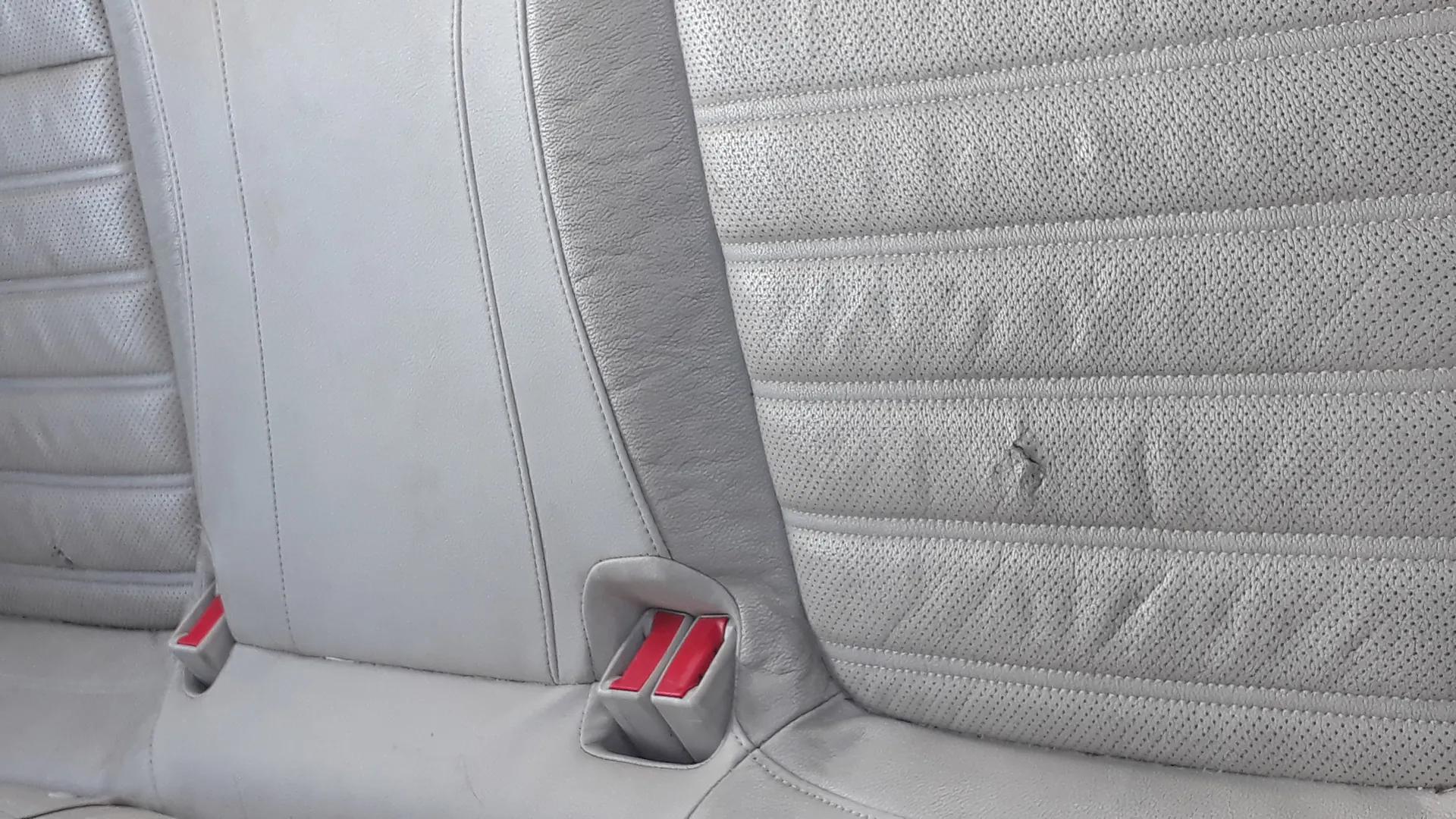 Zbliżenie na fragment jasnoszarego, perforowanego siedzenia samochodowego z widocznymi czerwonymi zatrzaskami pasów bezpieczeństwa. Siedzenie posiada poziome szwy i teksturę z drobnymi otworami. W prawej części obrazu widoczne jest niewielkie uszkodzenie materiału.