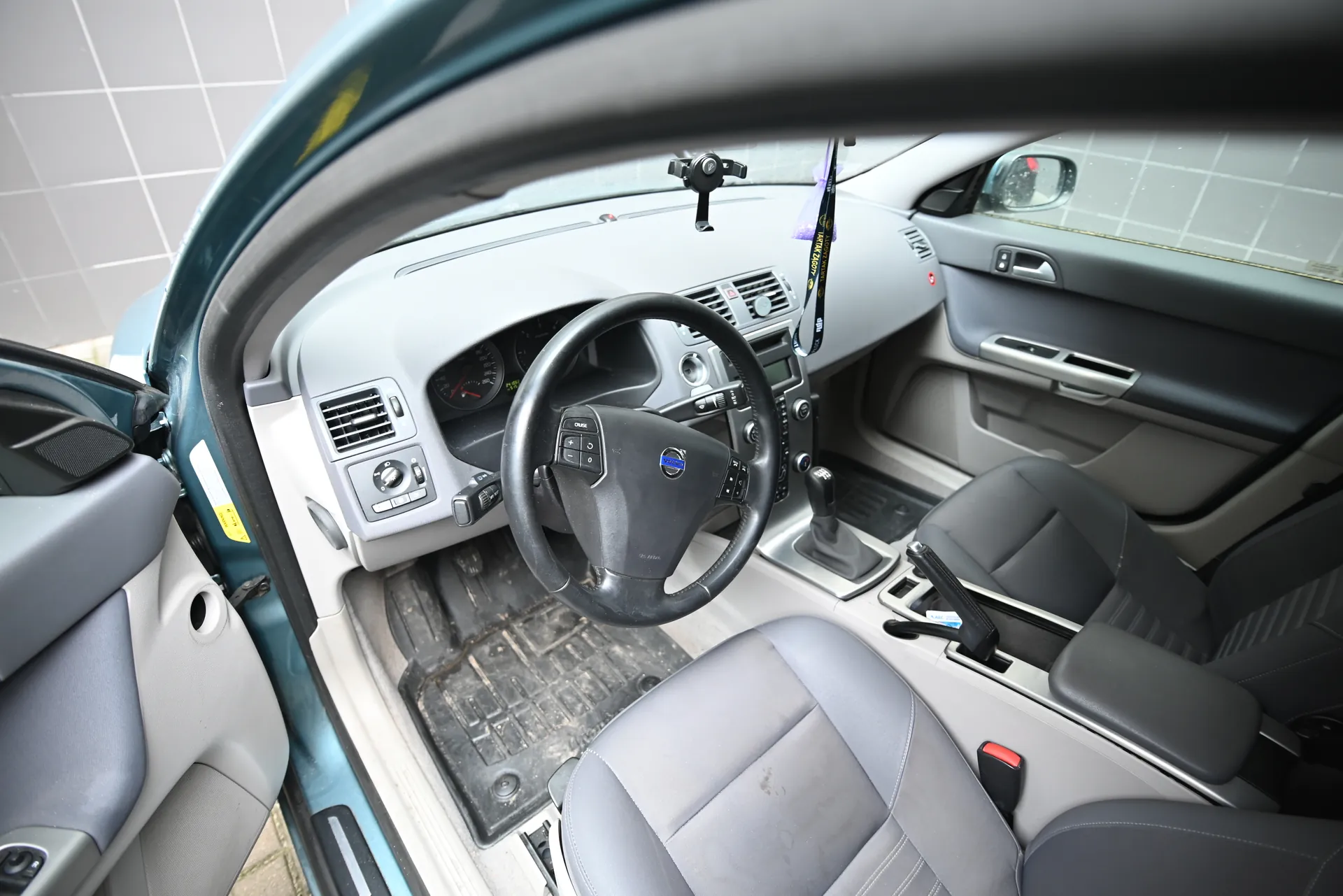 Wnętrze samochodu Volvo z otwartą lewą przodem drzwi. Widoczna kierownica z logo Volvo, deska rozdzielcza z licznikami, klimatyzacja, przyciski sterujące, dźwignia zmiany biegów oraz siedzenia w kolorze szaro-czarnym. Na desce rozdzielczej znajduje się czarny uchwyt na telefon. W drzwiach widoczne są żółte naklejki ostrzegawcze. Podłoga pokryta jest brudnymi czarnymi dywanikami. Poza samochodem widać szarą płytkową ścianę.