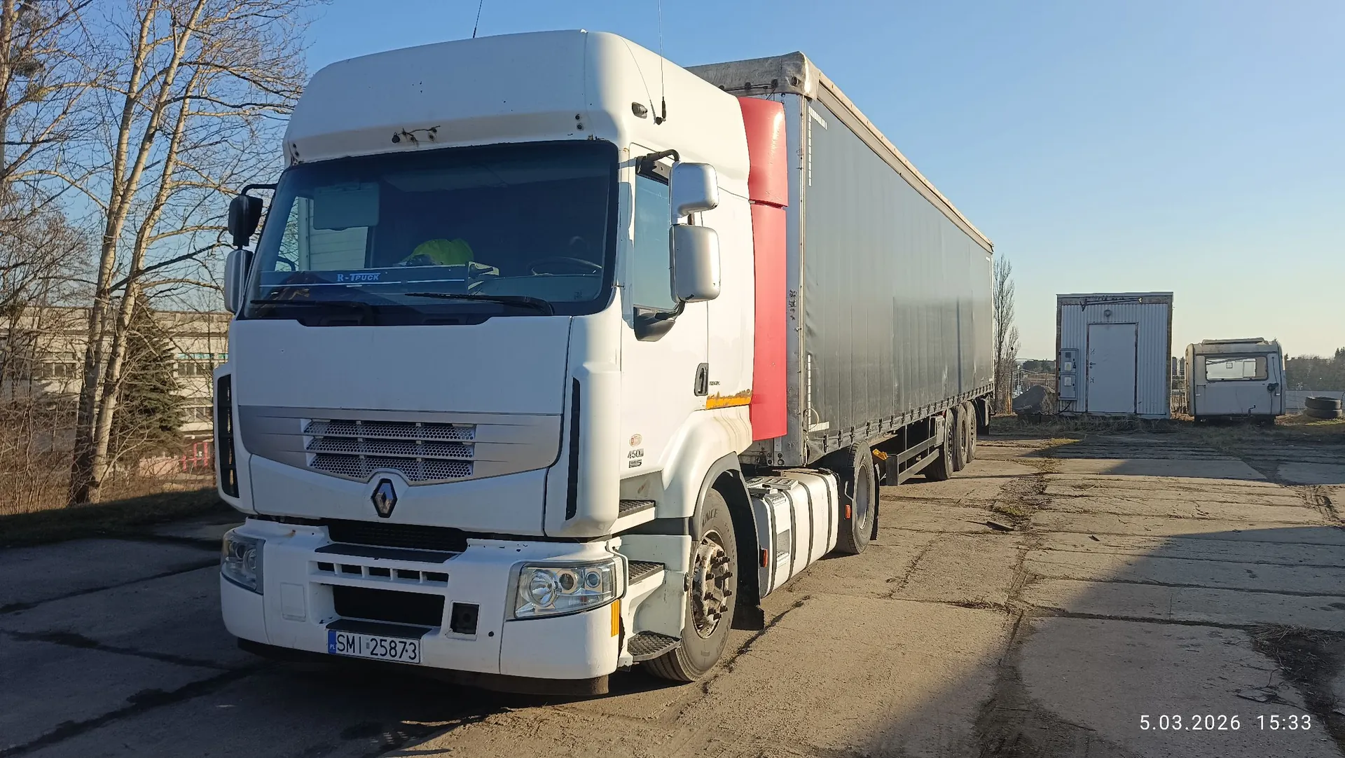 Biały ciągnik siodłowy marki Renault z naczepą typu kurtynowego w kolorze szarym z czerwonym pasem z przodu. Pojazd zaparkowany jest na betonowym placu. Na przednim zderzaku widoczna jest tablica rejestracyjna o numerze SMI 25873. W tle widać bezlistne drzewa, budynek oraz mały biały kontener. W prawym dolnym rogu znajduje się data i godzina: 5.03.2026 15:33.