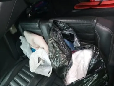 Wnętrze samochodu z czarnymi skórzonymi siedzeniami. Na tylnym siedzeniu znajdują się czarna torba plastikowa, biała torba plastikowa, jasny materiał lub ubranie oraz czerwony przedmiot po prawej stronie. Widoczne są również fragmenty drzwi i oparć siedzeń.