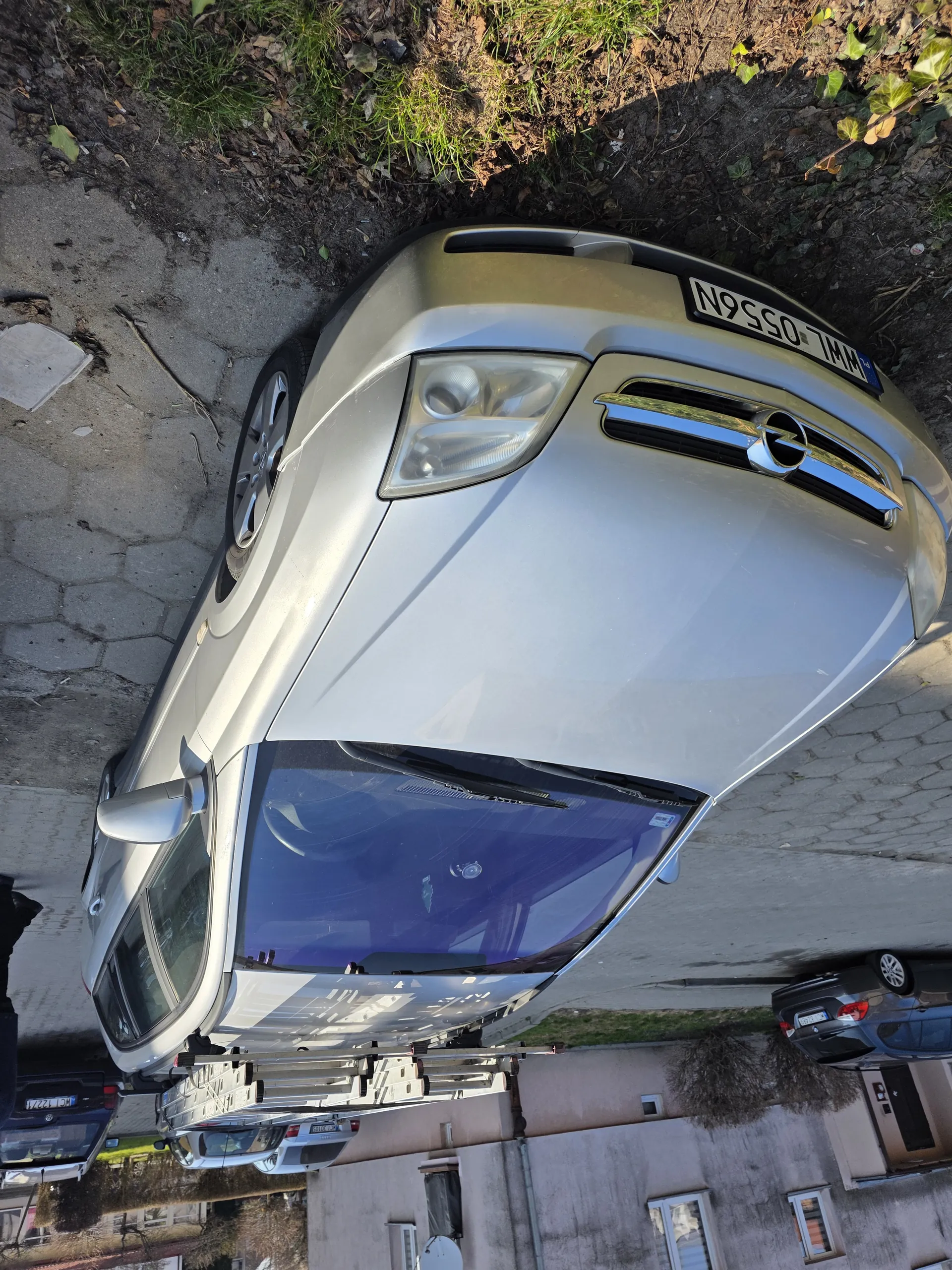 Srebrny samochód marki Opel zaparkowany na brukowanej ulicy. Na dachu widoczna drabina. Tablica rejestracyjna z numerem WHL 0556N. W tle budynki z oknami, satelitą, inne zaparkowane pojazdy oraz obszar z trawą i suchymi liśćmi. Nawierzchnia ulicy składa się z sześciokątnych płyt.