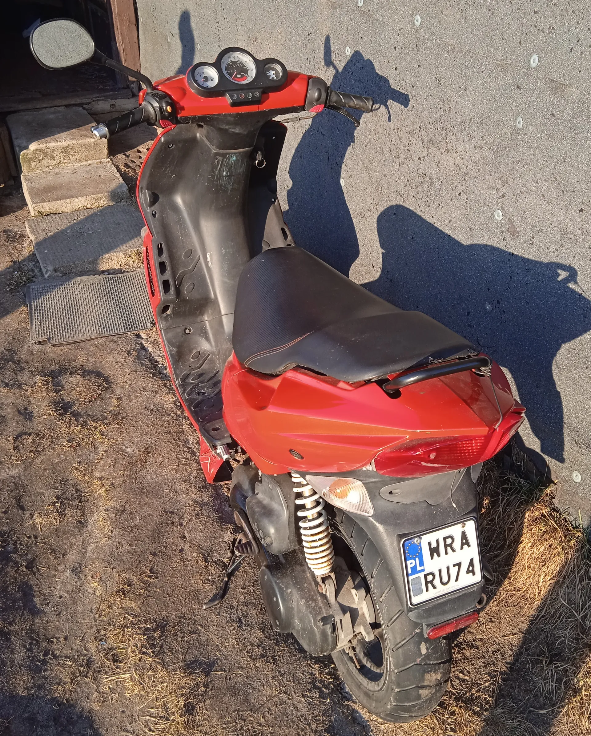 Czerwony skuter z czarnym siedzeniem i czarnymi elementami, zaparkowany na brudnej ziemi z suchą trawą obok asfaltowej nawierzchni. Widoczna tablica rejestracyjna z białym tłem, niebieskim pasem z napisem "PL" oraz czarnymi literami i cyframi "WRA RU74". W tle schody z betonowych stopni, wejście do budynku i cień skutera na ziemi. Skuter posiada lusterka, wskaźniki na desce rozdzielczej oraz tylną oś z widoczną sprężyną.