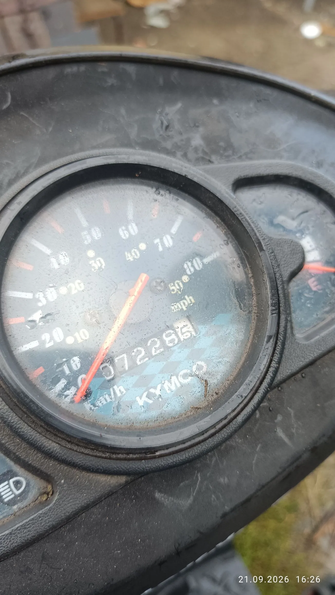 Zbliżenie na licznik prędkości skutera marki KYMCO. Tarcza licznika posiada dwie skale: zewnętrzną w mph (mile na godzinę) oraz wewnętrzną w km/h (kilometry na godzinę). Czerwona wskazówka wskazuje wartość bliską 0. Licznik kilometrów pokazuje wartość 072286. Szybka licznika jest zarysowana i zabrudzona. W prawym dolnym rogu zdjęcia widnieje data i godzina: 21.09.2026 16:26.