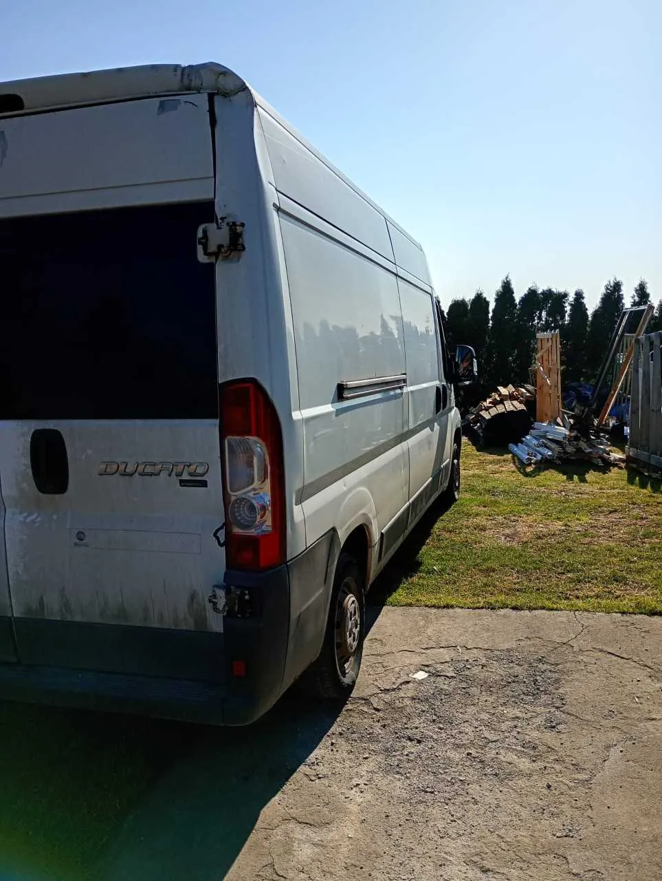 Biały furgon marki Ducato zaparkowany na betonowej powierzchni. Widoczne są zanieczyszczenia na tylnym panelu, ciemne tylne okno oraz czerwone światła. W tle trawa, stosy materiałów budowlanych, drzewa i jasne niebo.