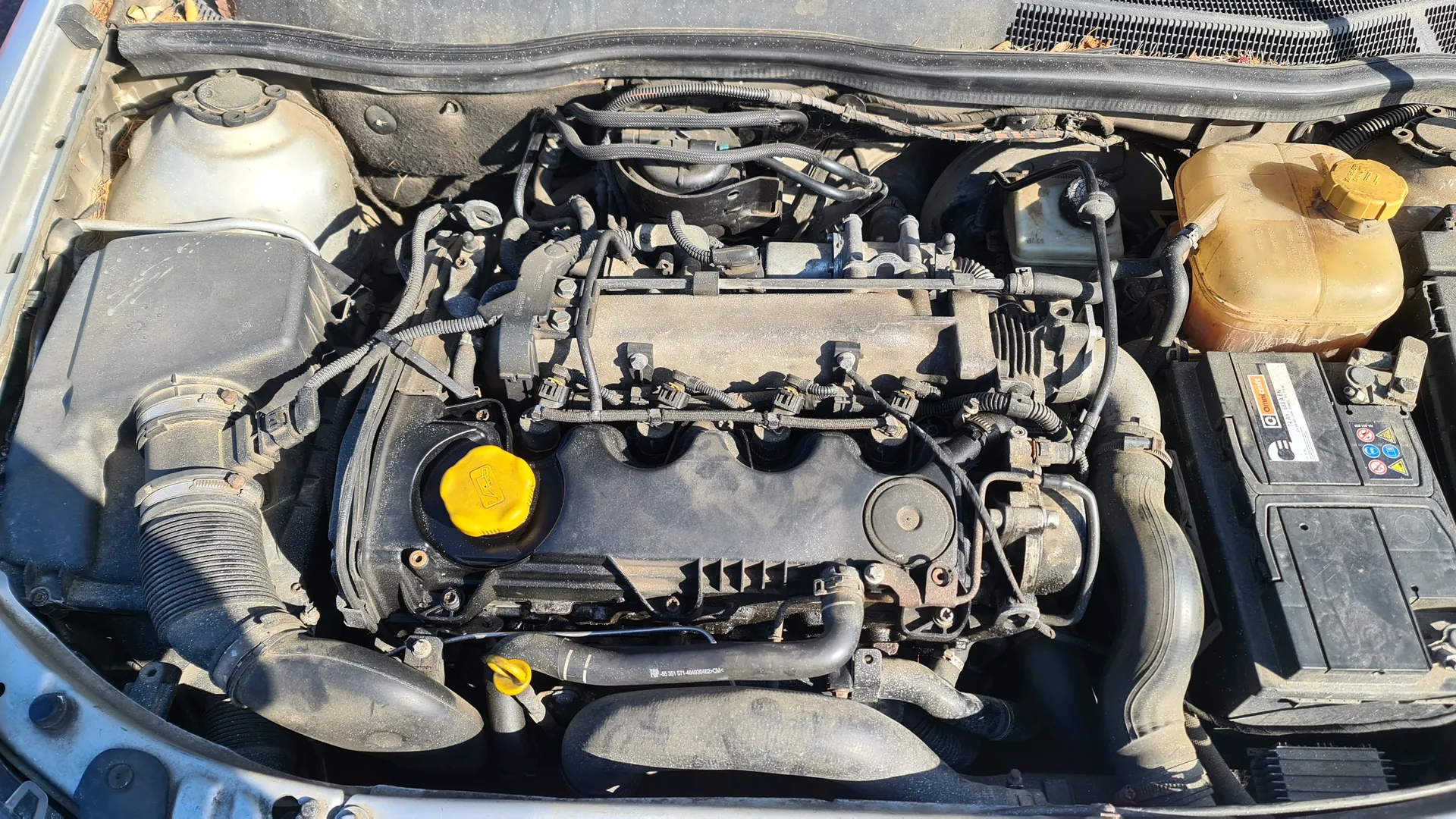 Widok na komorę silnika samochodu z otwartą maską. W centrum znajduje się czarny silnik z żółtym korkiem na zbiorniku oleju. Po lewej stronie srebrny zbiornik chłodniczy, po prawej żółtawy zbiornik płynu hamulcowego z żółtym korkiem. W prawym dolnym rogu czarna bateria z etykietami i symbolami. Wszystkie elementy pokryte są kurzem. Widoczne są czarne przewody, węże oraz różne części mechaniczne.