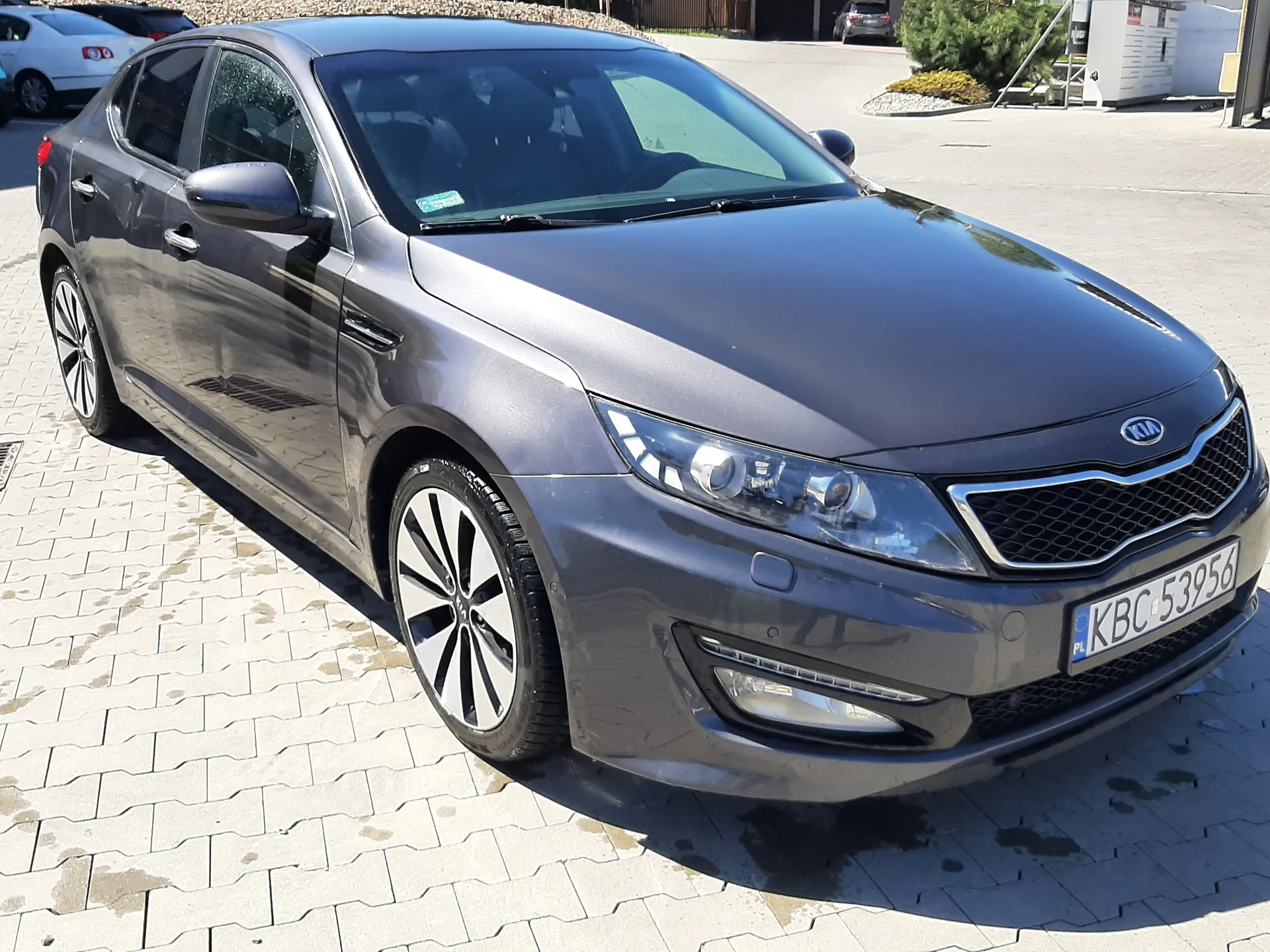 Srebrny samochód marki Kia, model sedan, zaparkowany na brukowanej powierzchni. Widoczna tablica rejestracyjna z polskim kodem PL i numerem KBC 53956. Na przedniej szybie naklejka. W tle inne pojazdy, budynki, krzewy i droga. Oświetlenie słoneczne, cienie wskazują na jasny dzień.