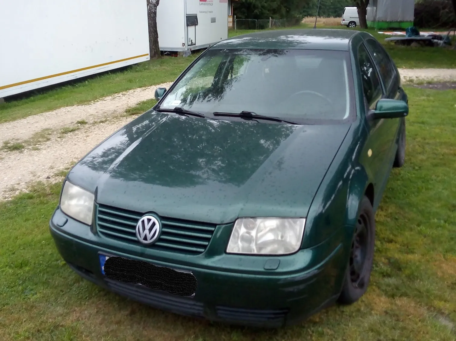 Ciemnozielony samochód marki Volkswagen zaparkowany na trawie obok żwirowej drogi. Widoczny jest przód pojazdu z logo VW na grillu, reflektory oraz zamaskowana tablica rejestracyjna. W tle znajdują się białe przyczepy i fragmenty zieleni.