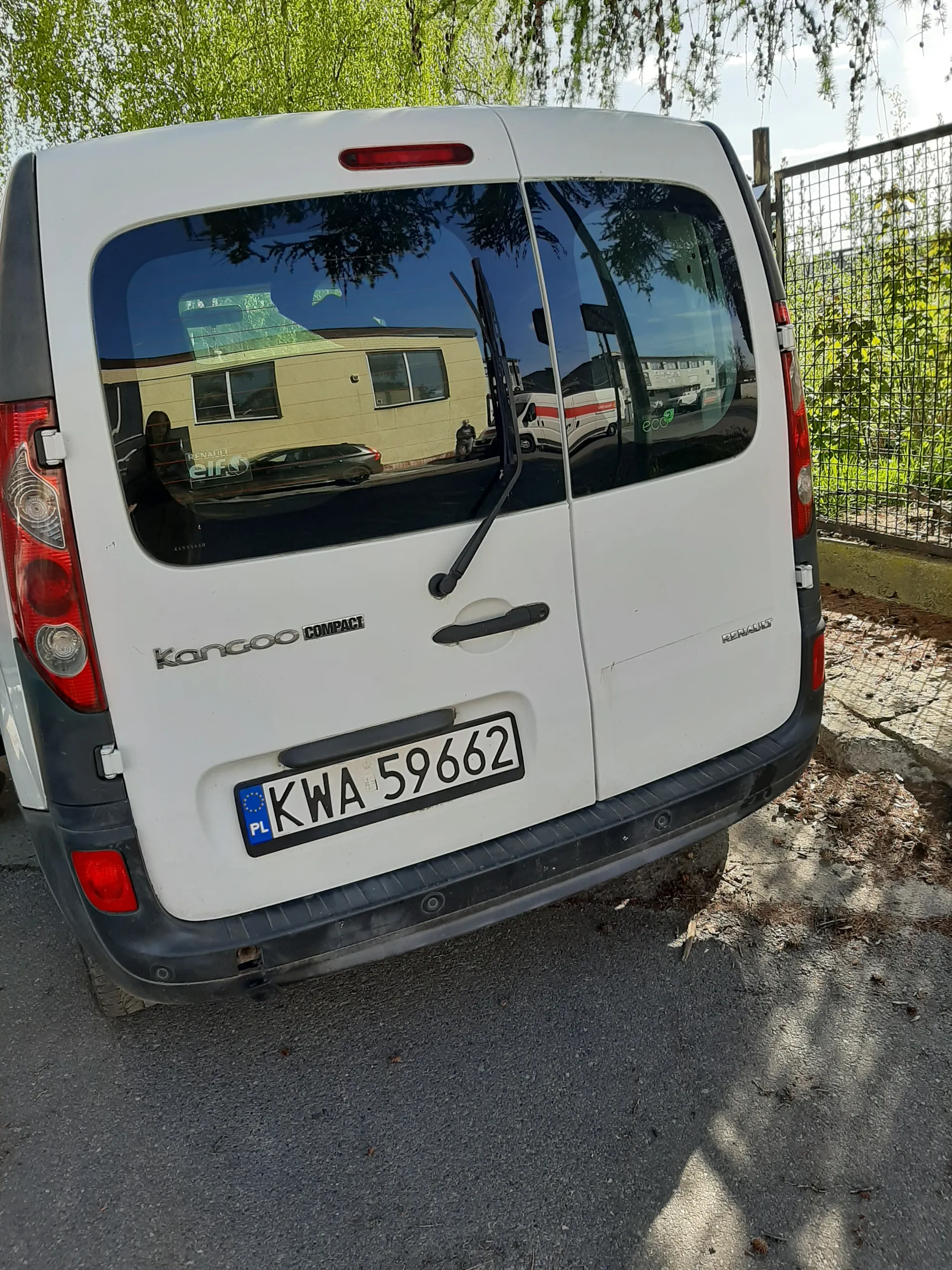 Biały samochód dostawczy Renault Kangoo Compact widziany od tyłu. Tablica rejestracyjna KWA 59662 PL umieszczona pionowo z niebieskim pasem z napisem PL i symbolem UE. W tle siatka, zielone drzewa, kamienista ściana oraz asfaltowa powierzchnia. W szybie tylnej odbicie budynku, innych pojazdów i drzew.