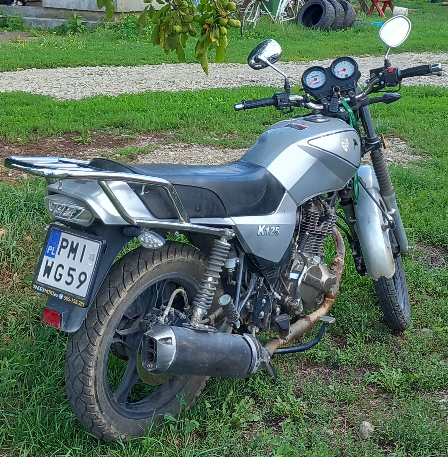 Srebrny motocykl marki K125 zaparkowany na trawie. Widoczny jest tył i bok pojazdu, z tablicą rejestracyjną o numerze PMI WG59. Motocykl posiada czarną kanapę, chromowany bagażnik oraz czarną rurę wydechową.
