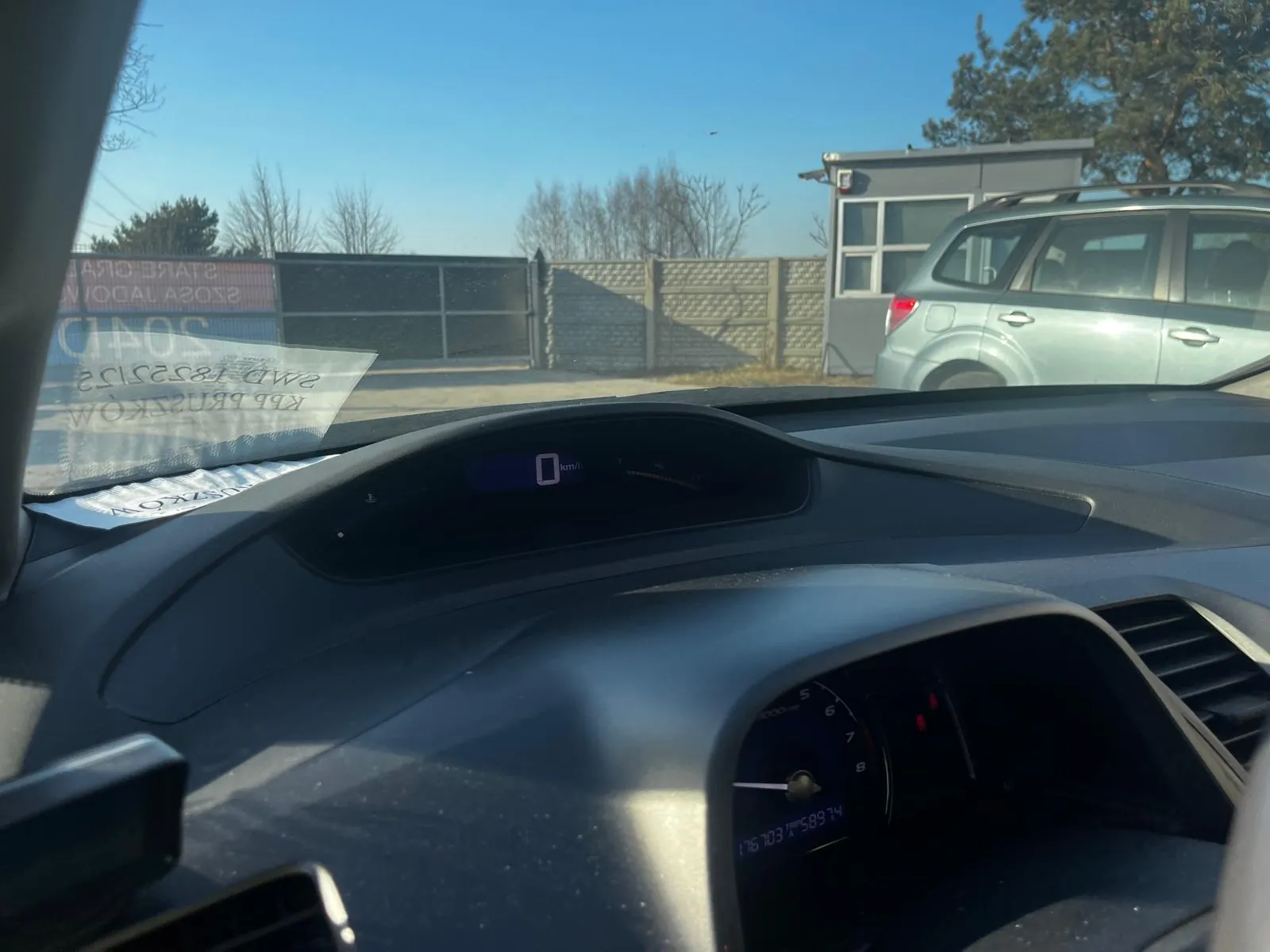 Zdjęcie z wnętrza samochodu przedstawiające deskę rozdzielczą z prędkościomierzem wskazującym 0 km/h i cyfrowym wyświetlaczem. Poza przednią szybą widoczny srebrny SUV, ogrodzenie, niewielki budynek, drzewa oraz jasne niebo. Na przedniej szybie znajduje się naklejka z częściowo widocznym tekstem.