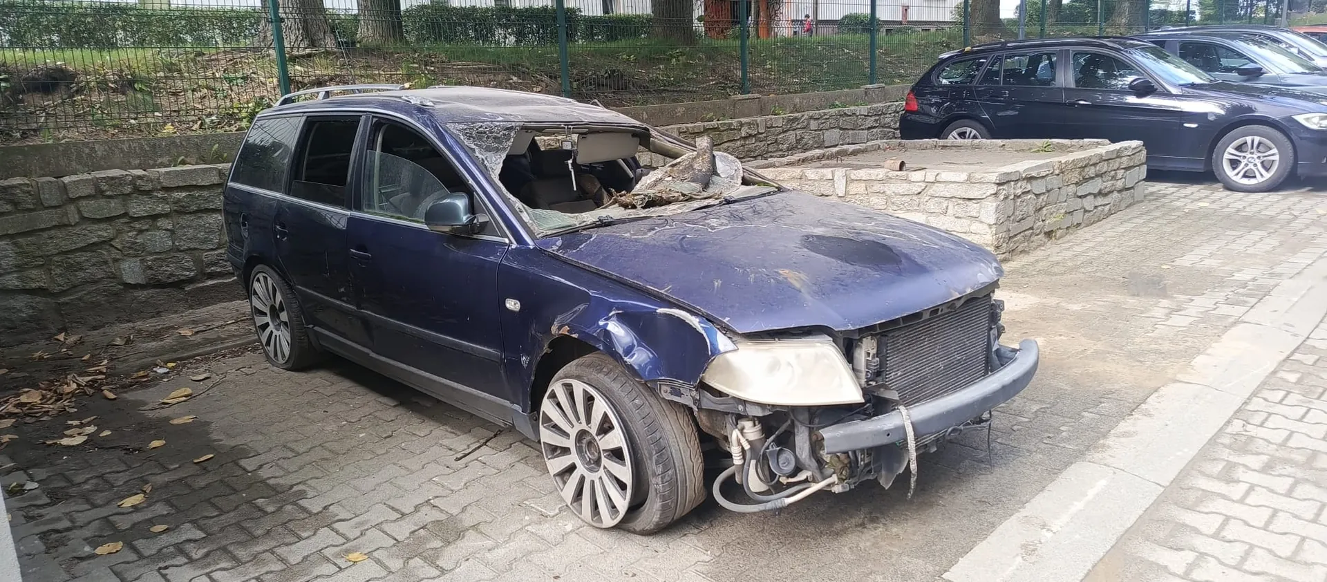 Granatowy samochód typu kombi z poważnymi uszkodzeniami przodu i dachu. Przednia szyba jest całkowicie rozbita, dach jest zapadnięty, brakuje przedniego zderzaka i części maski, a prawa przednia opona jest całkowicie rozszczelniona i spłaszczona. Auto stoi na kostce brukowej obok niskiego murka z kamienia. W tle widoczny jest inny, nieuszkodzony ciemny samochód oraz zielone drzewa i ogrodzenie.