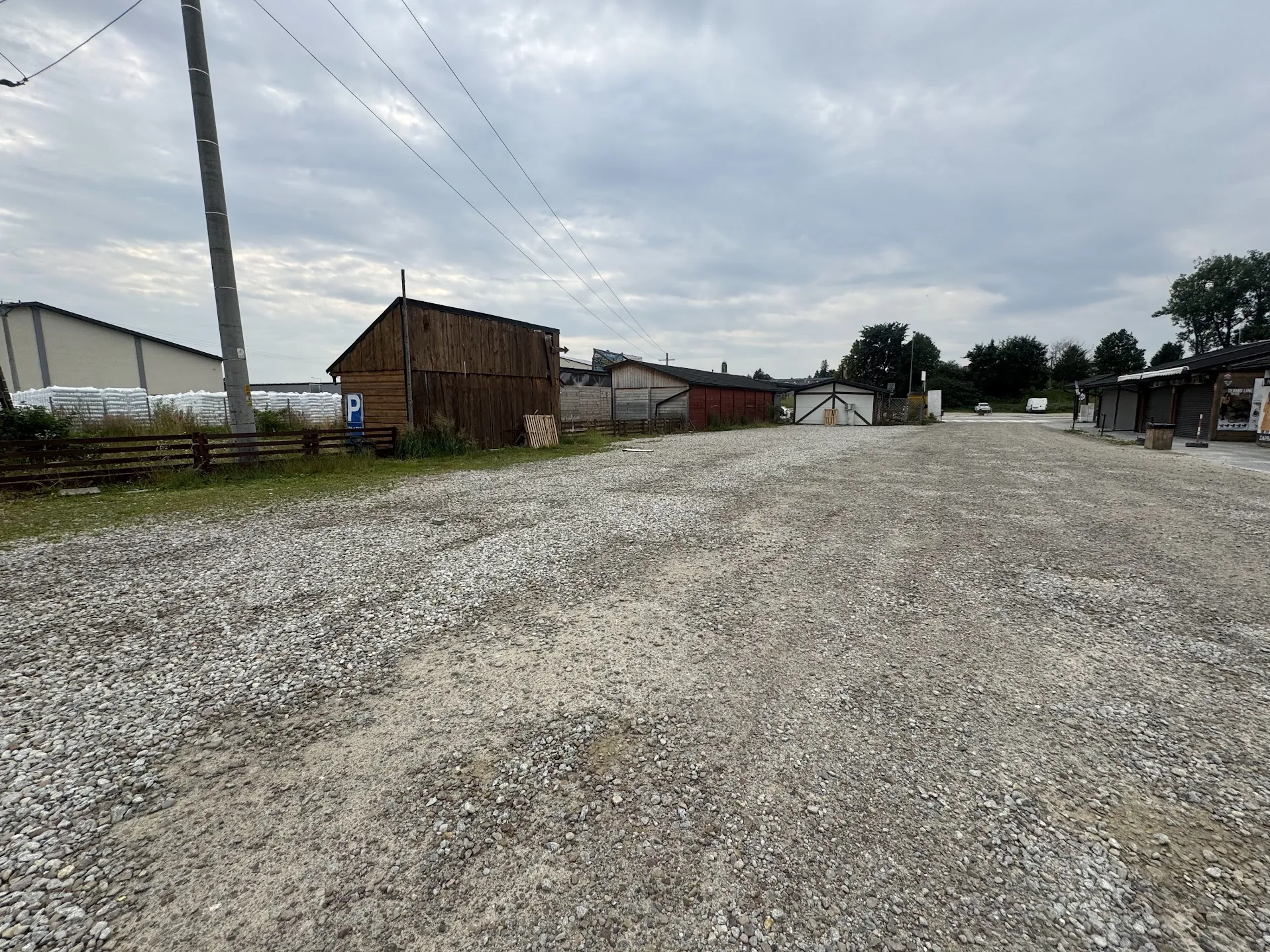 Szeroki widok na żwirowy plac przed grupą niskich budynków gospodarczych i magazynowych. Po lewej stronie znajduje się drewniany płot, za którym stoi ciemnobrązowy drewniany budynek oraz jasny budynek z białymi elementami. Widoczny jest niebieski znak parkingowy z literą 'P'. W tle widać inne zabudowania, drzewa oraz zachmurzone niebo. Po lewej stronie kadru przebiega pionowo betonowy słup energetyczny.