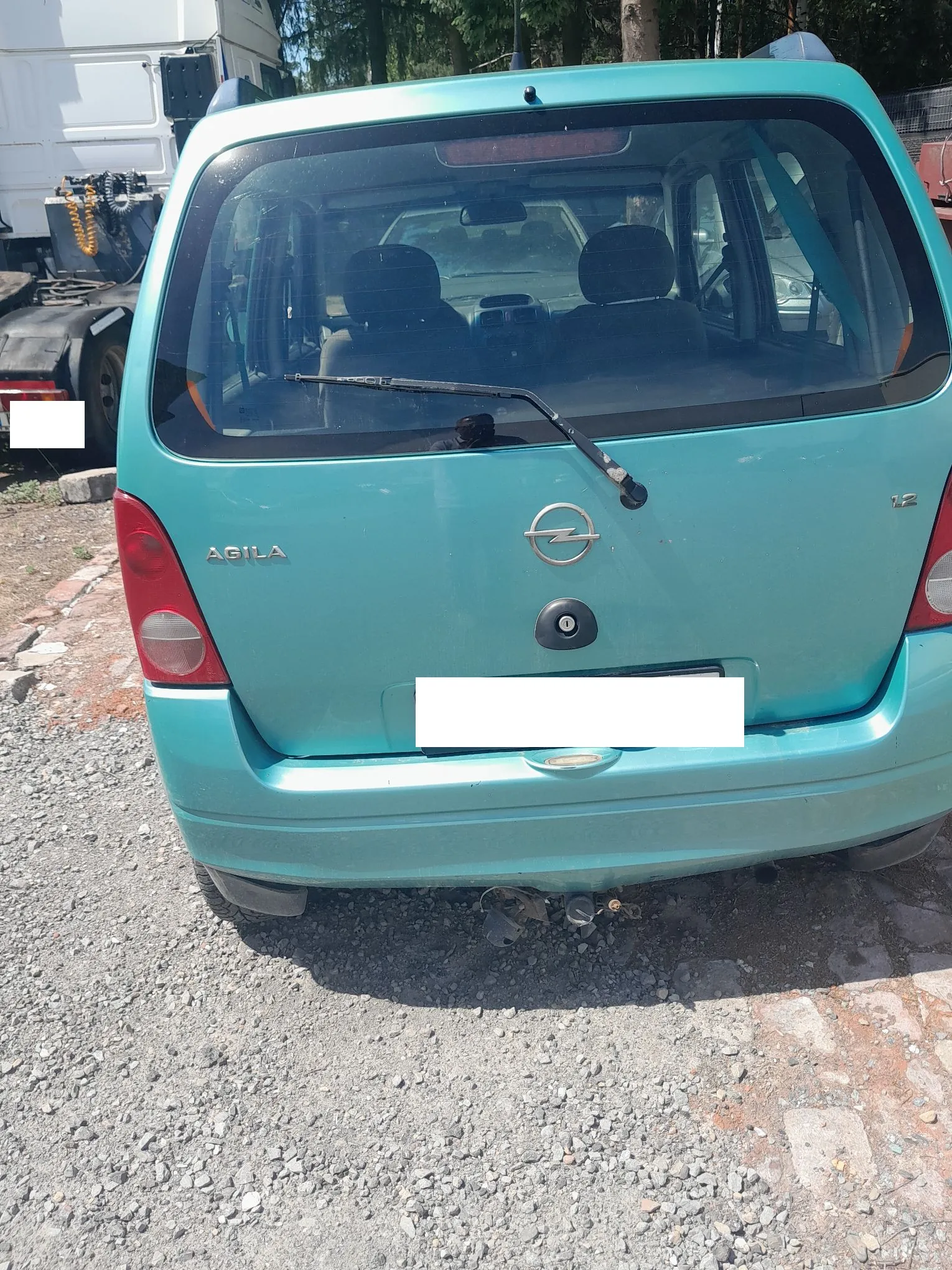Tylny widok turkusowego samochodu marki Opel Agila. Na klapie bagażnika widoczne jest logo Opela, napis 'AGILA' po lewej stronie oraz oznaczenie '1.2' po prawej. Tablica rejestracyjna jest zakryta białym prostokątem. Samochód posiada hak holowniczy i jest zaparkowany na żwirowym podłożu.