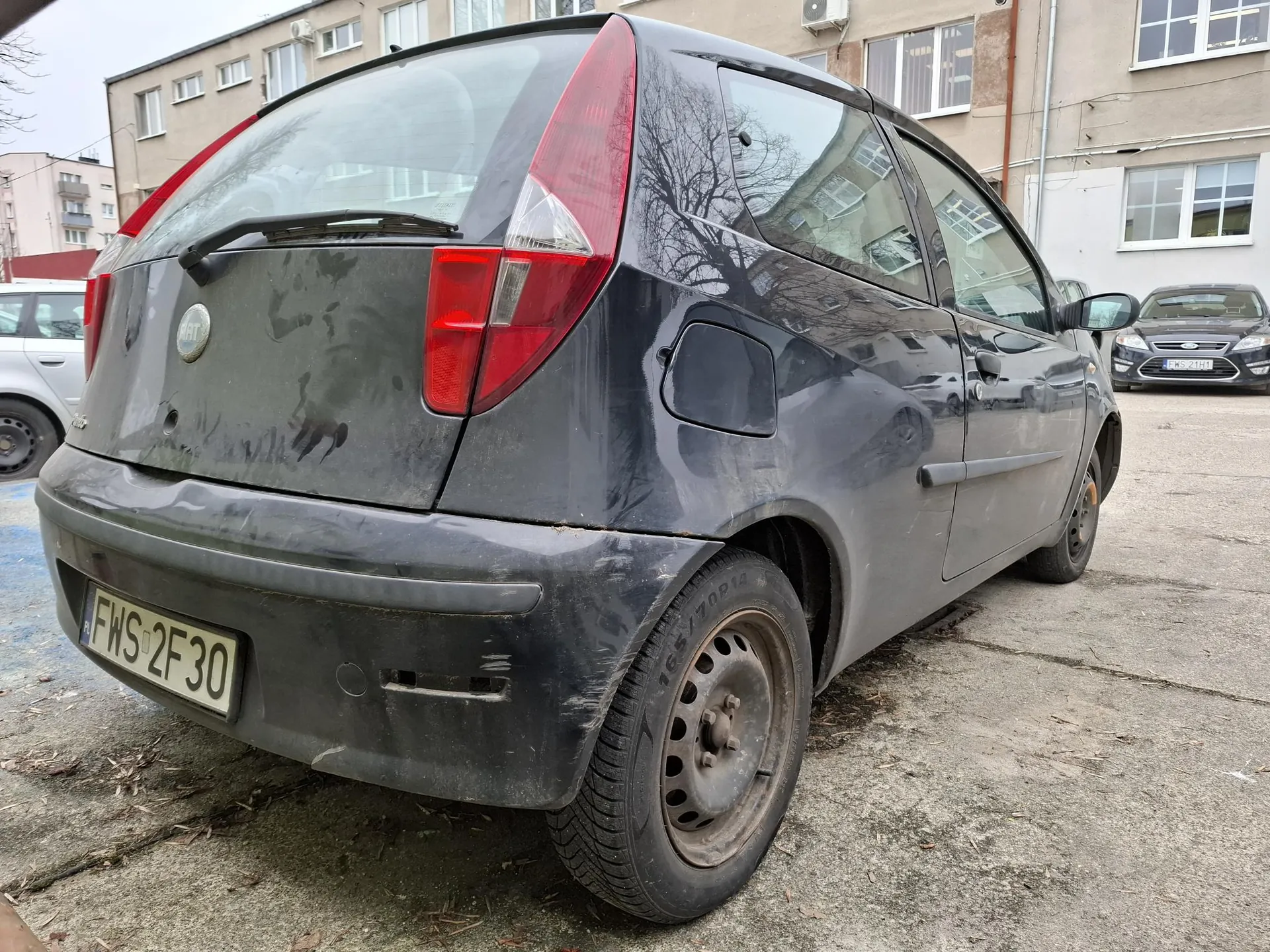 Czarny samochód Fiat z tyłu, widoczna tablica rejestracyjna FWS 2F30. Samochód jest zabrudzony, z widocznymi zadrapaniami. W tle budynki mieszkalne, inne zaparkowane pojazdy (srebrny i czarny samochód) oraz betonowy parking. Na tylnej szybie refleksy drzew i budynków.