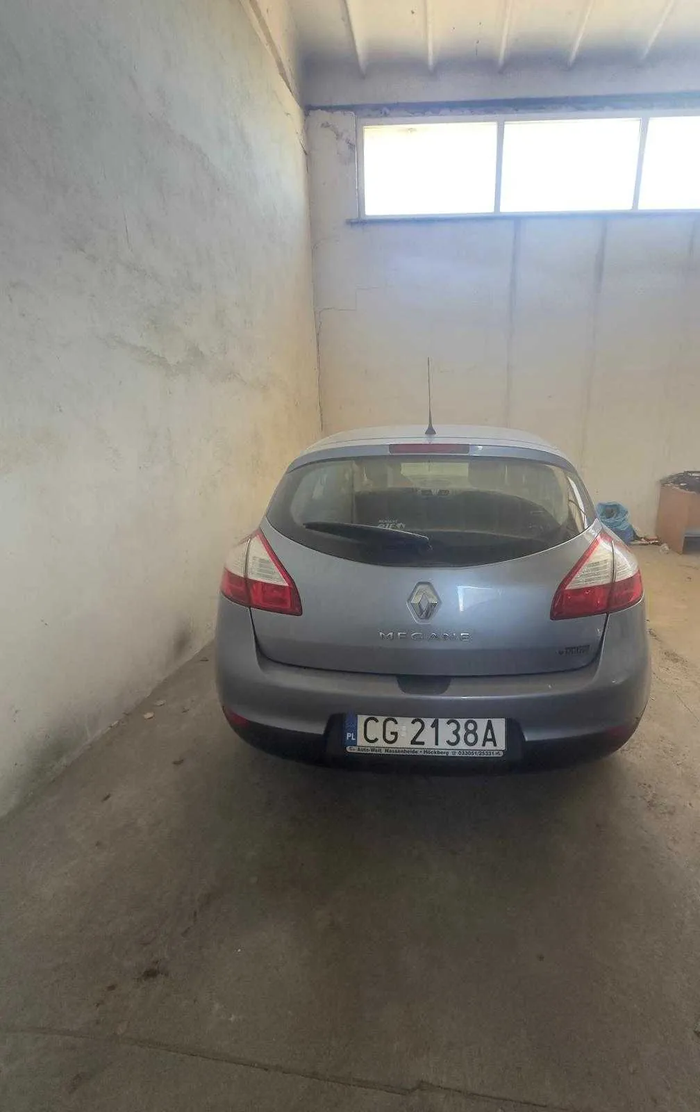 Srebrny samochód Renault Megane widziany od tyłu, zaparkowany w garażu z betonowymi ścianami. Na tylnej szybie widoczny wycieraczka, a na bagażniku logo marki i napis "MEGANE". Tablica rejestracyjna z polskim kodem PL i numerem CG 2138A. W tle okno z trzema segmentami oraz na prawo od samochodu niebieski worek i karton na betonowej podłodze.