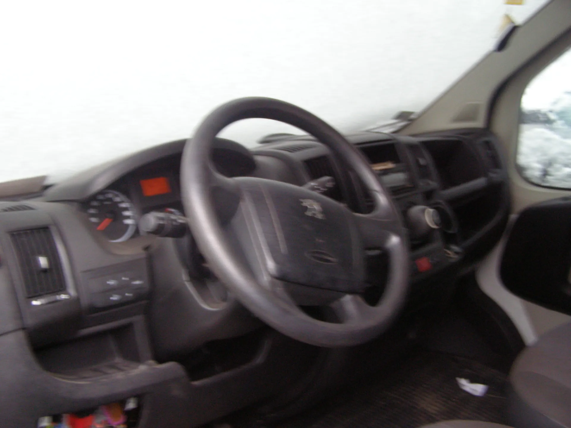 Wnętrze samochodu z ciemnoszarym panelem deski rozdzielczej, kierownicą z logo, prędkościomierzem, pomarańczowym wyświetlaczem, klimatyzacją, przyciskami sterowania, szarymi siedzeniami oraz oknem po prawej stronie ukazującym zasneżony krajobraz na zewnątrz.