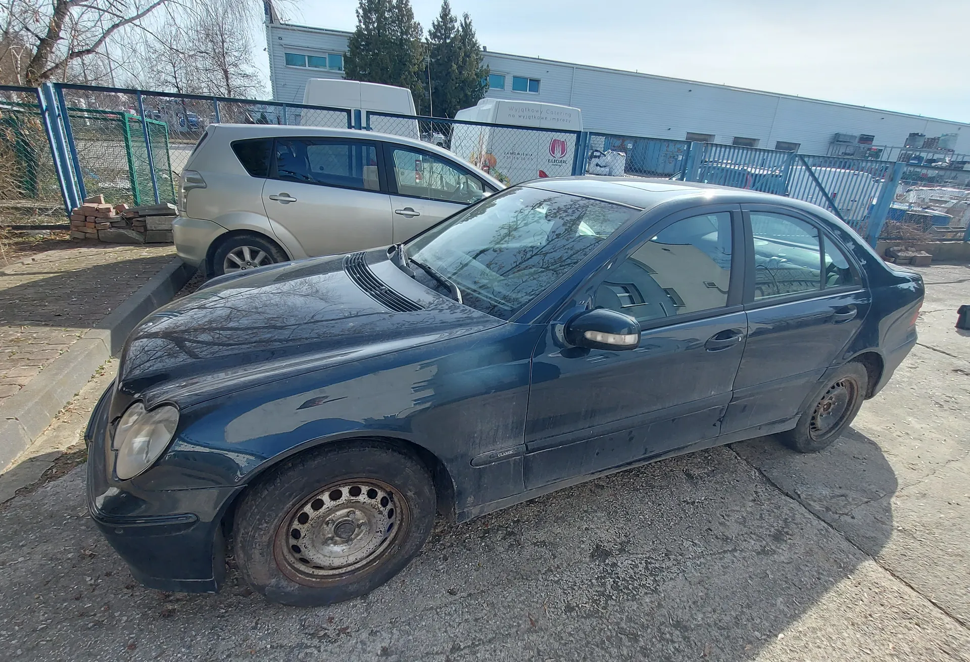 Ciemnoniebieski sedan osobowy w stanie zużycia, zaparkowany na betonowej powierzchni. Samochód ma zabrudzoną karoserię, zarysowane felgi i opony z widoczną korozją. Za nim stoi srebrny samochód typu hatchback. W tle widoczne są niebieskie ogrodzenie, budynek przemysłowy, drzewa bez liści oraz biały furgon. Niebo jest jasne, a całość znajduje się na otwartym placu parkingowym.