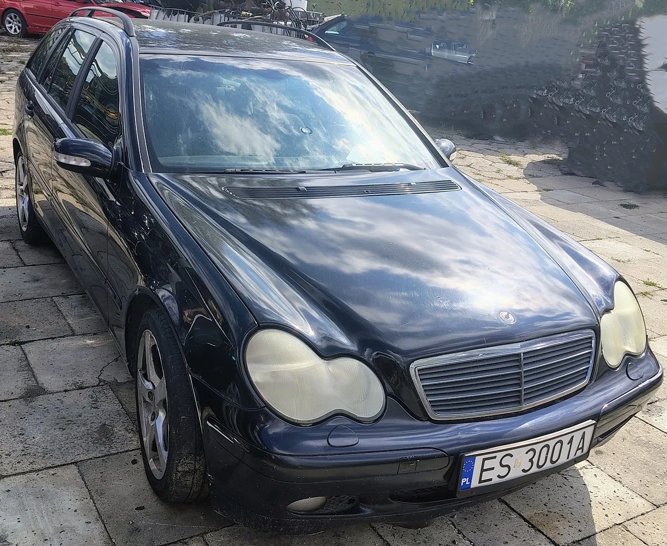 Czarny samochód osobowy marki Mercedes-Benz w nadwoziu kombi, zaparkowany na betonowym placu. Pojazd ma widoczne zmatowiałe reflektory przednie oraz polską tablicę rejestracyjną o numerze ES 3001A.