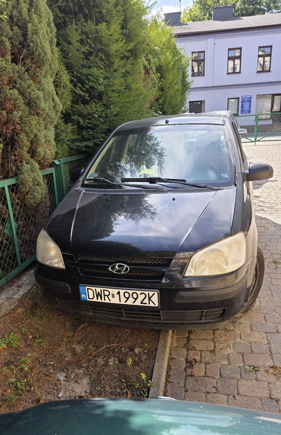 Czarny samochód marki Hyundai zaparkowany na kostce brukowej obok zielonego ogrodzenia i wysokich, gęstych krzewów. Samochód ma polską tablicę rejestracyjną o numerze DWR 1992K. W tle widoczny jest jasnofioletowy, dwupiętrowy budynek z oknami.