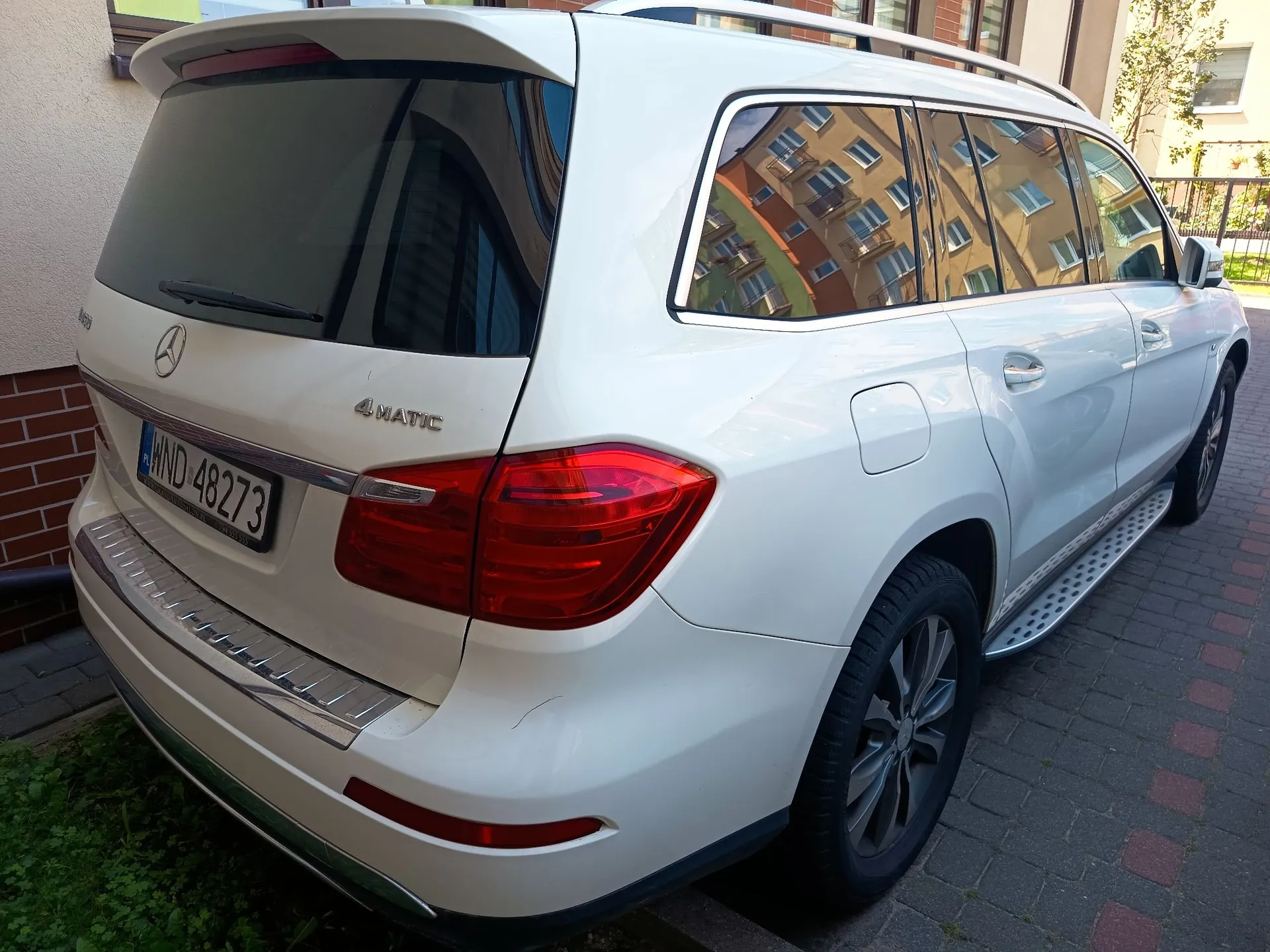 Biały samochód marki Mercedes-Benz klasy GL w wersji 4MATIC, zaparkowany na kostce brukowej. Widoczny jest tył i prawy bok pojazdu. Samochód posiada tablicę rejestracyjną o numerze WND 48273. W bocznych szybach odbijają się kolorowe fasady budynków mieszkalnych.