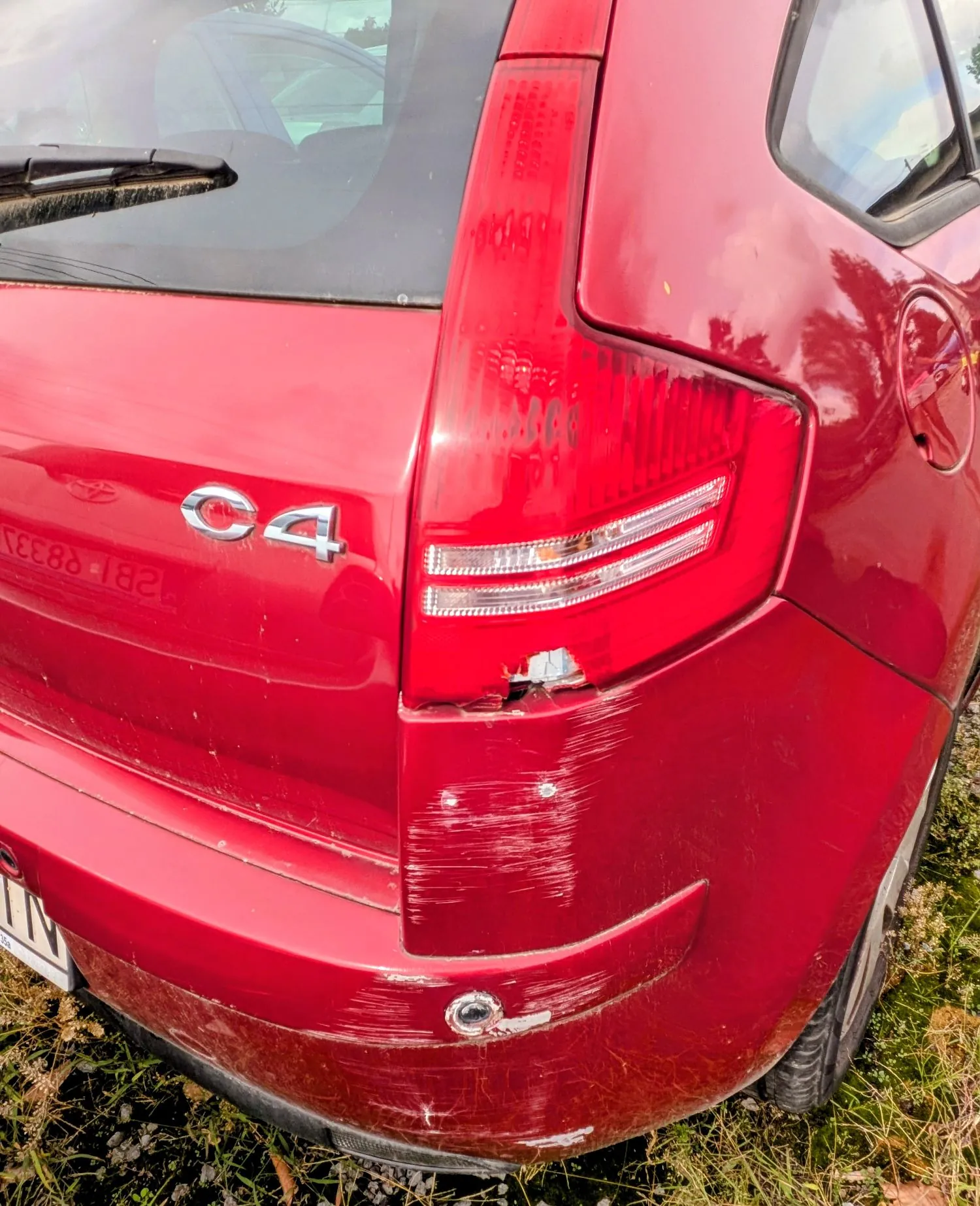 Zbliżenie na tył czerwonego samochodu marki Citroën C4. Widoczny jest napis 'C4' oraz tylna lampa. Na zderzaku i karoserii w okolicy lampy znajdują się liczne zarysowania, otarcia oraz pęknięcie w dolnej części klosza lampy.