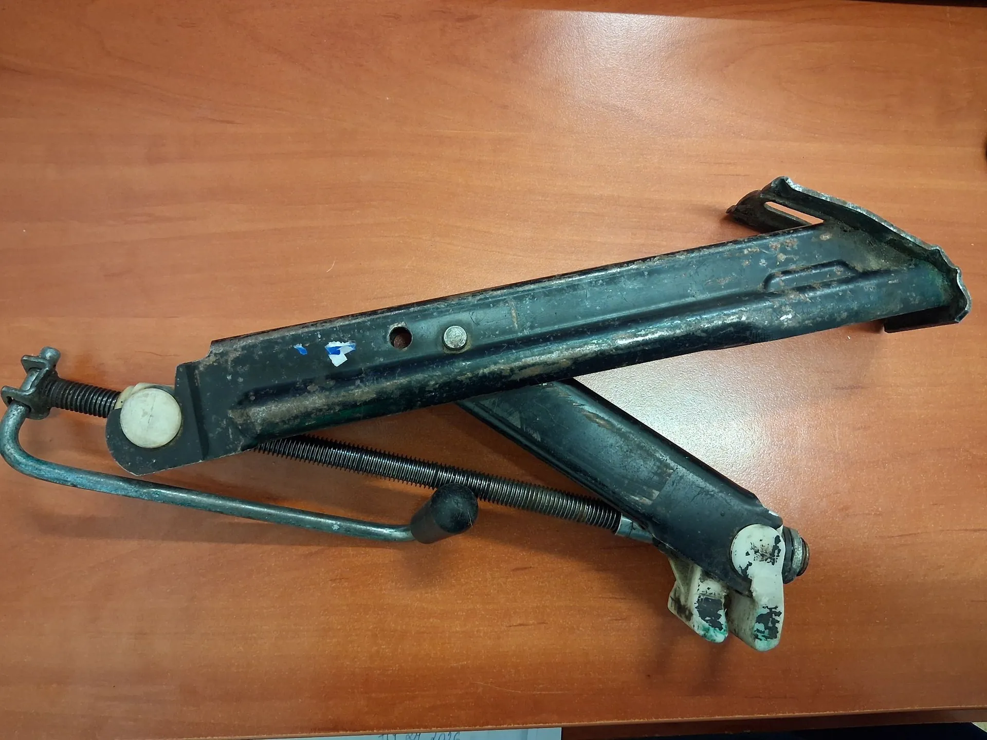 Metalowy, czarny mechanizm podnośnika szyby samochodowej leżący na brązowym, drewnianym blacie. Element składa się z długiej szyny, ramienia poprzecznego oraz metalowej sprężyny i prowadnicy.