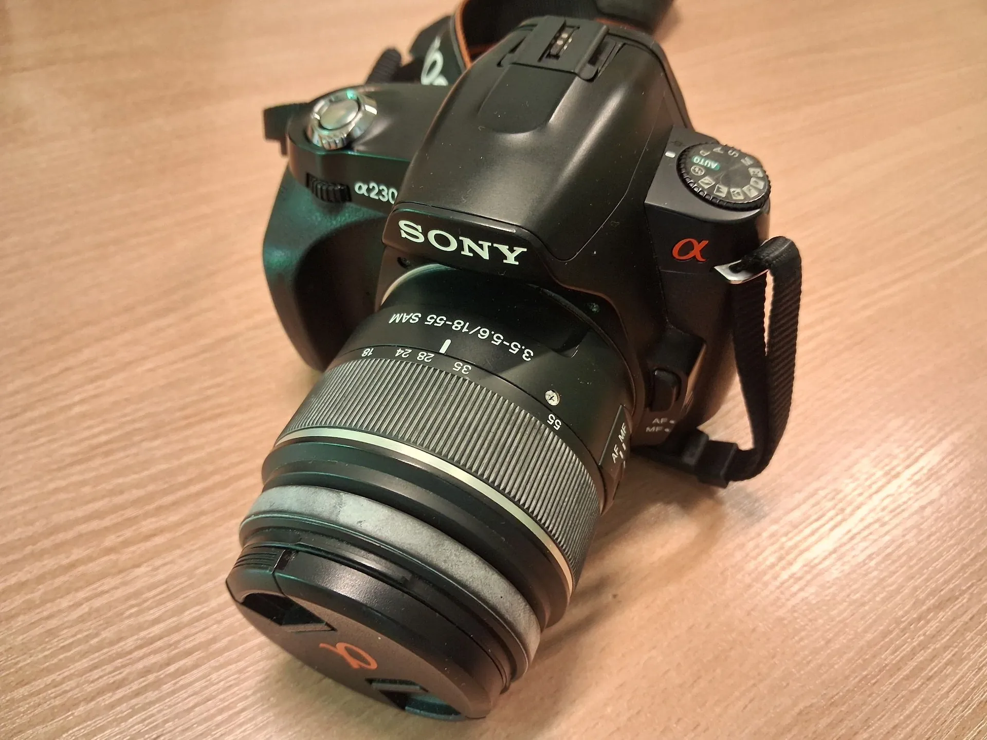 Czarny aparat fotograficzny Sony z obiektywem, leżący na jasnym, drewnianym blacie. Na korpusie widoczne są napisy 'SONY', 'α230' oraz czerwony symbol 'α'. Na obiektywie znajduje się napis '3.5-5.6/18-55 SAM'. Aparat jest wyposażony w czarny pasek na szyję.