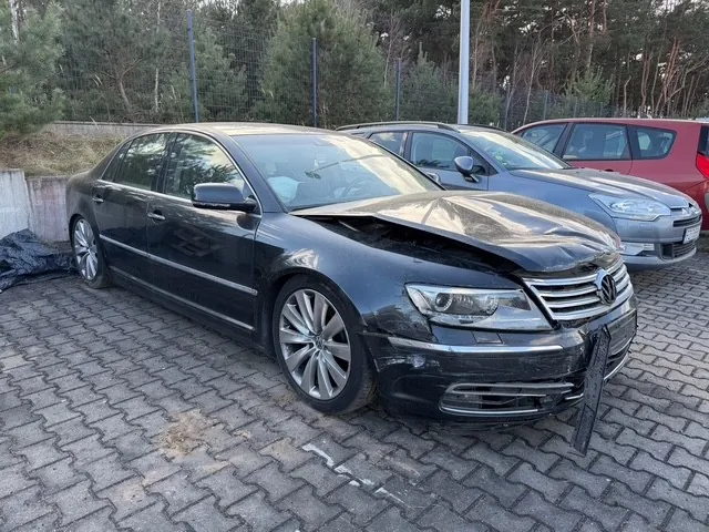 Czarny samochód osobowy marki Volkswagen z uszkodzonym przodem, zaparkowany na brukowanym placu. Widoczne są zgniecione maski i zderzak. Za nim stoją srebrny i czerwony samochód. Tło stanowi ogrodzenie, drzewa i zarośla. Na ziemi leży czarna torba.