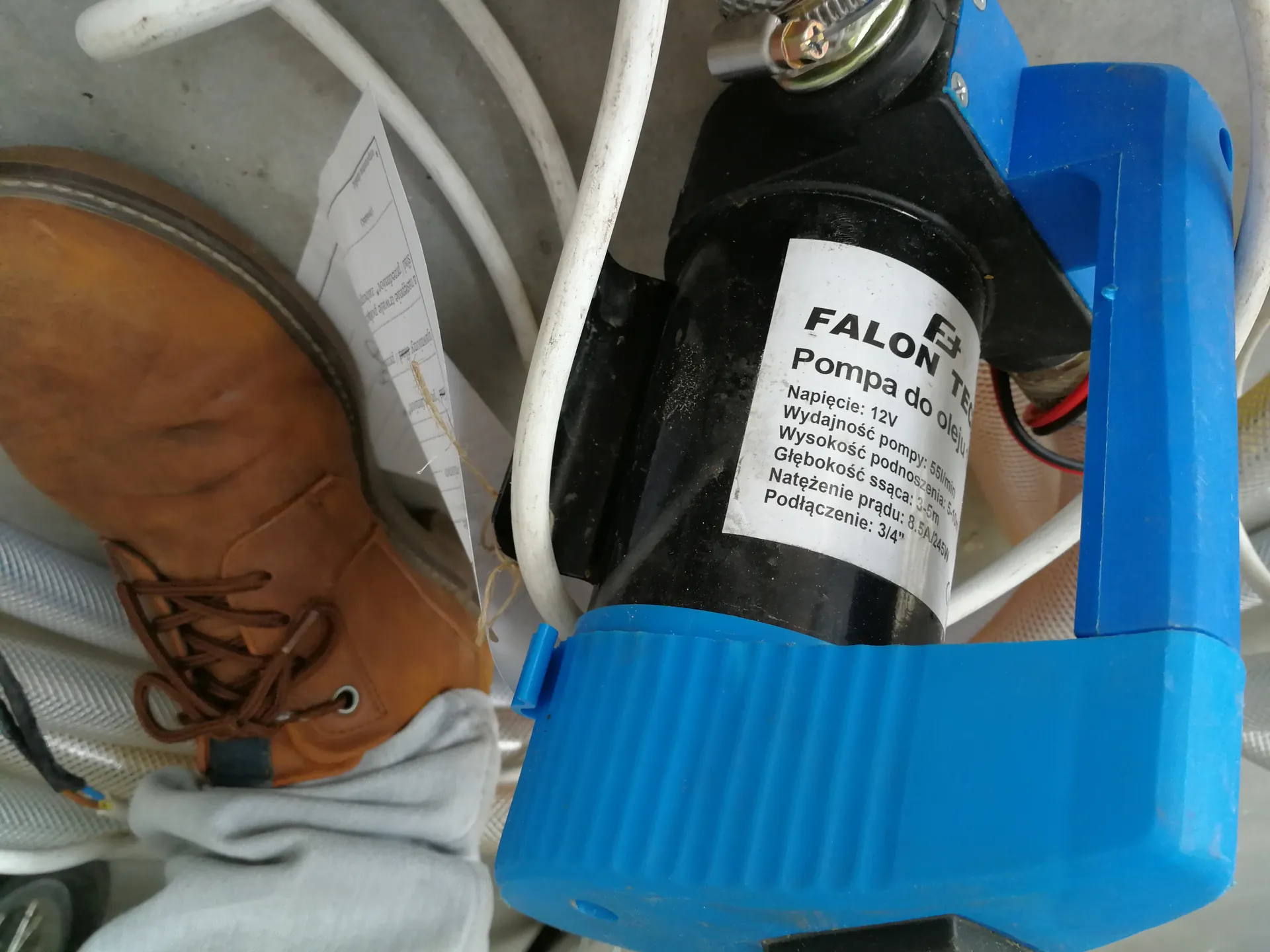 Niebiesko-czarna pompa z białą etykietą zawierającą tekst: 'FALON TECH Pompa do oleju, Napięcie: 12V, Wydajność pompy: 5l/min, Wysokość podnoszenia: 5-10m, Głębokość ssania: 3-5m, Natężenie prądu: 8.5A/12V, Połączenie: 3/4"'. Obok pompy brązowy but roboczy z szarymi spodniami, białe przewody i fragment papieru z tekstem.